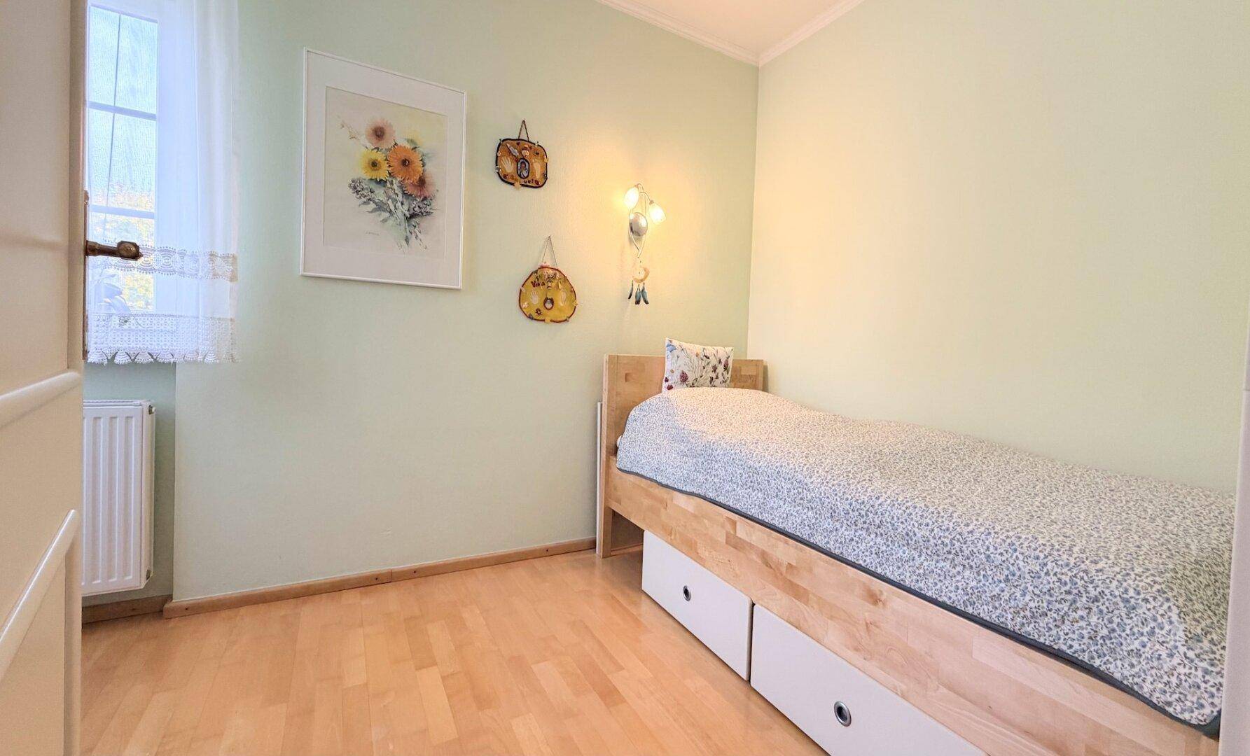 Helles Kinder- oder Gästezimmer mit Einzelbett, Holzboden und Fenster, grüne Wände.