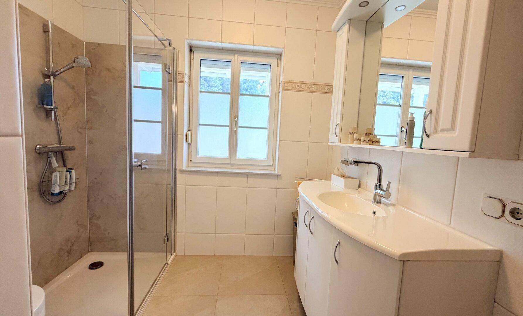 Modernes Badezimmer mit Dusche, Einzelwaschbecken und Fenster, hell gefliest.