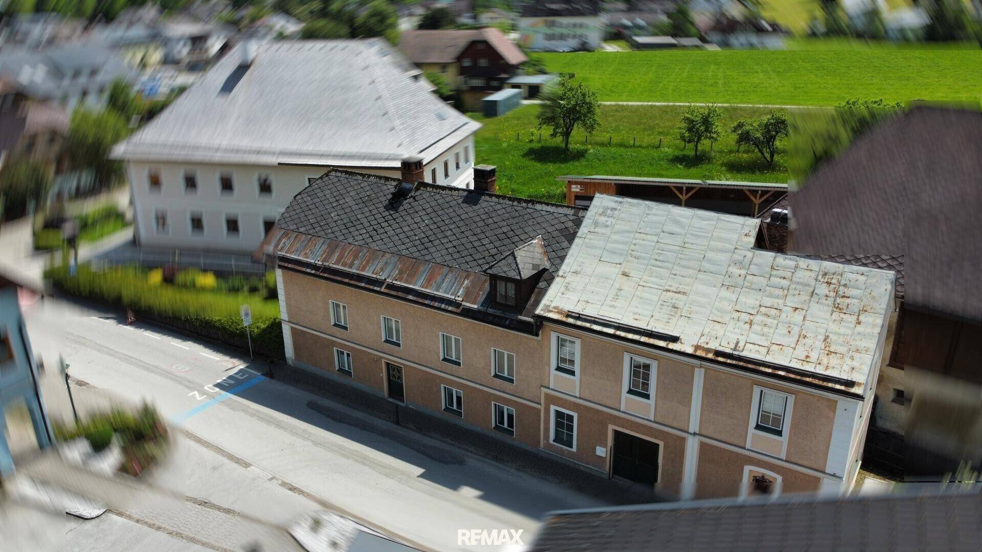 Luftaufnahme eines Gebäudes an einer Straße mit angrenzenden Grünflächen und weiteren Häusern.