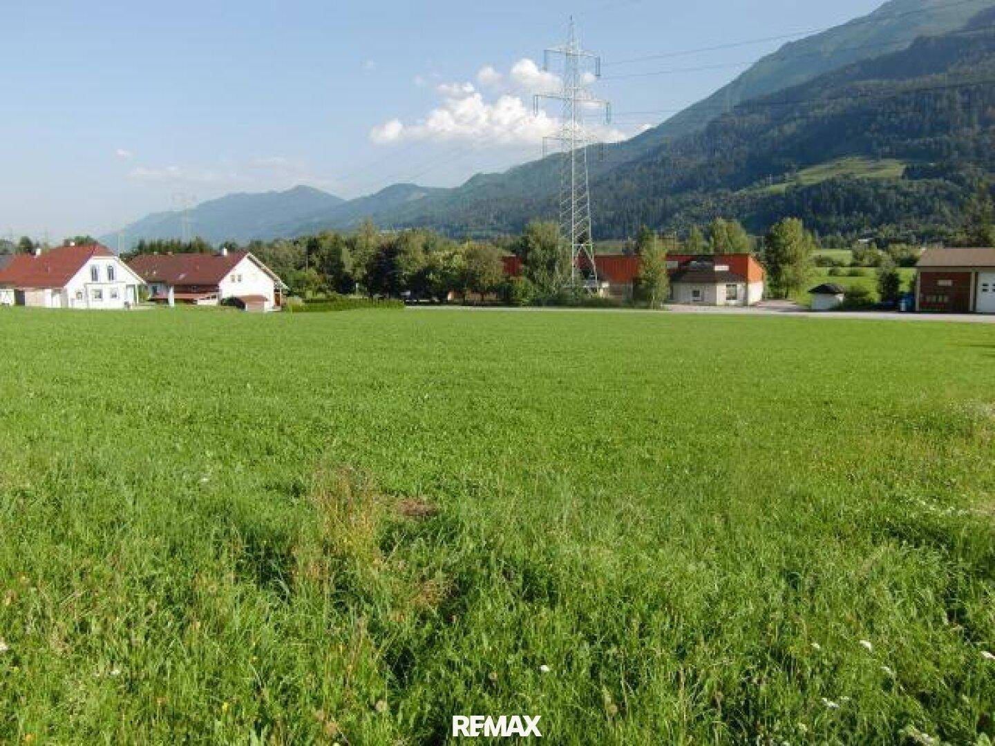 Weitläufige grüne Wiese mit Bergblick und vereinzelten Häusern in der Ferne unter blauem Himmel.
