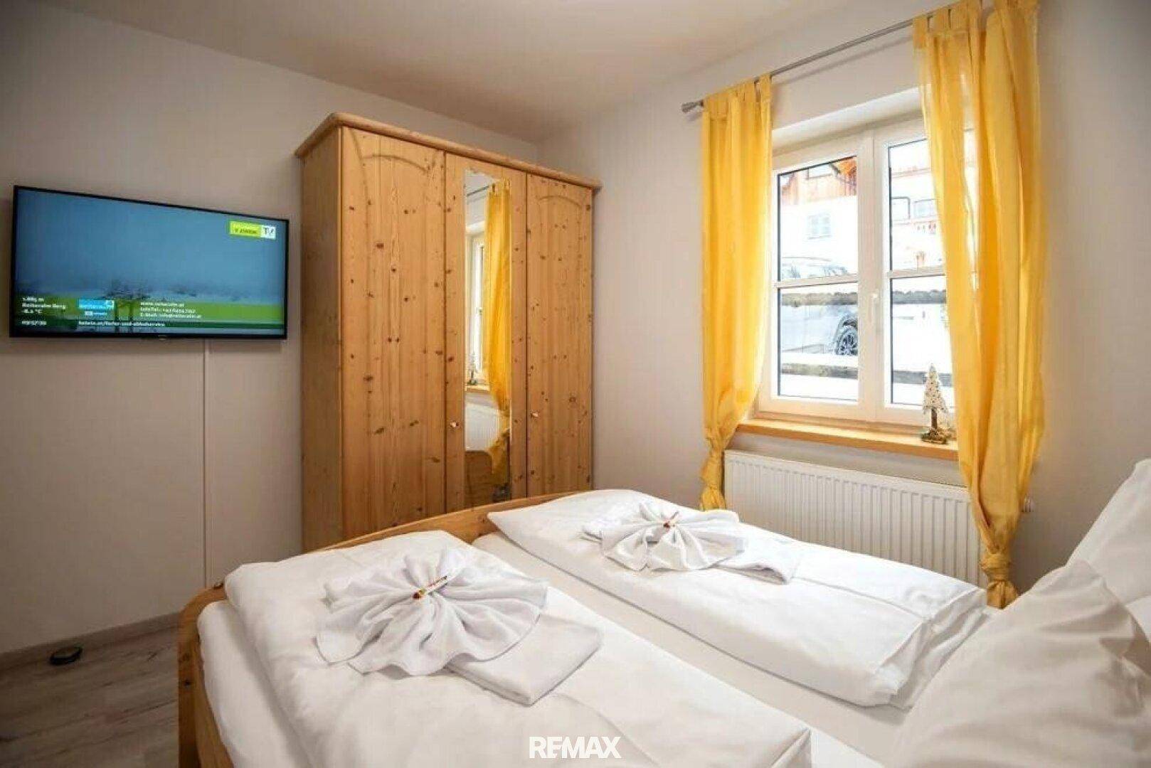 Gemütliches Schlafzimmer mit zwei Einzelbetten, Holzschrank, gelben Vorhängen und Wandfernseher.