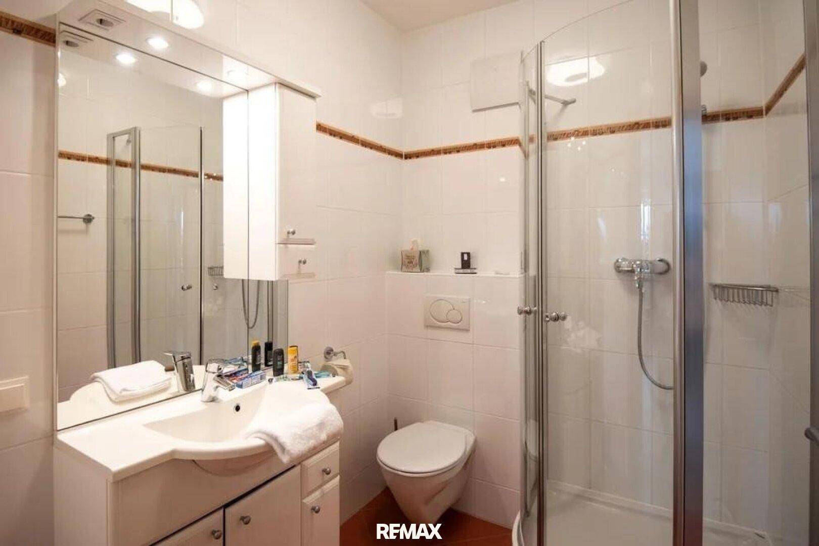 Helles Badezimmer mit weißen Fliesen, brauner Bordüre, Waschtisch, Toilette und Duschkabine.