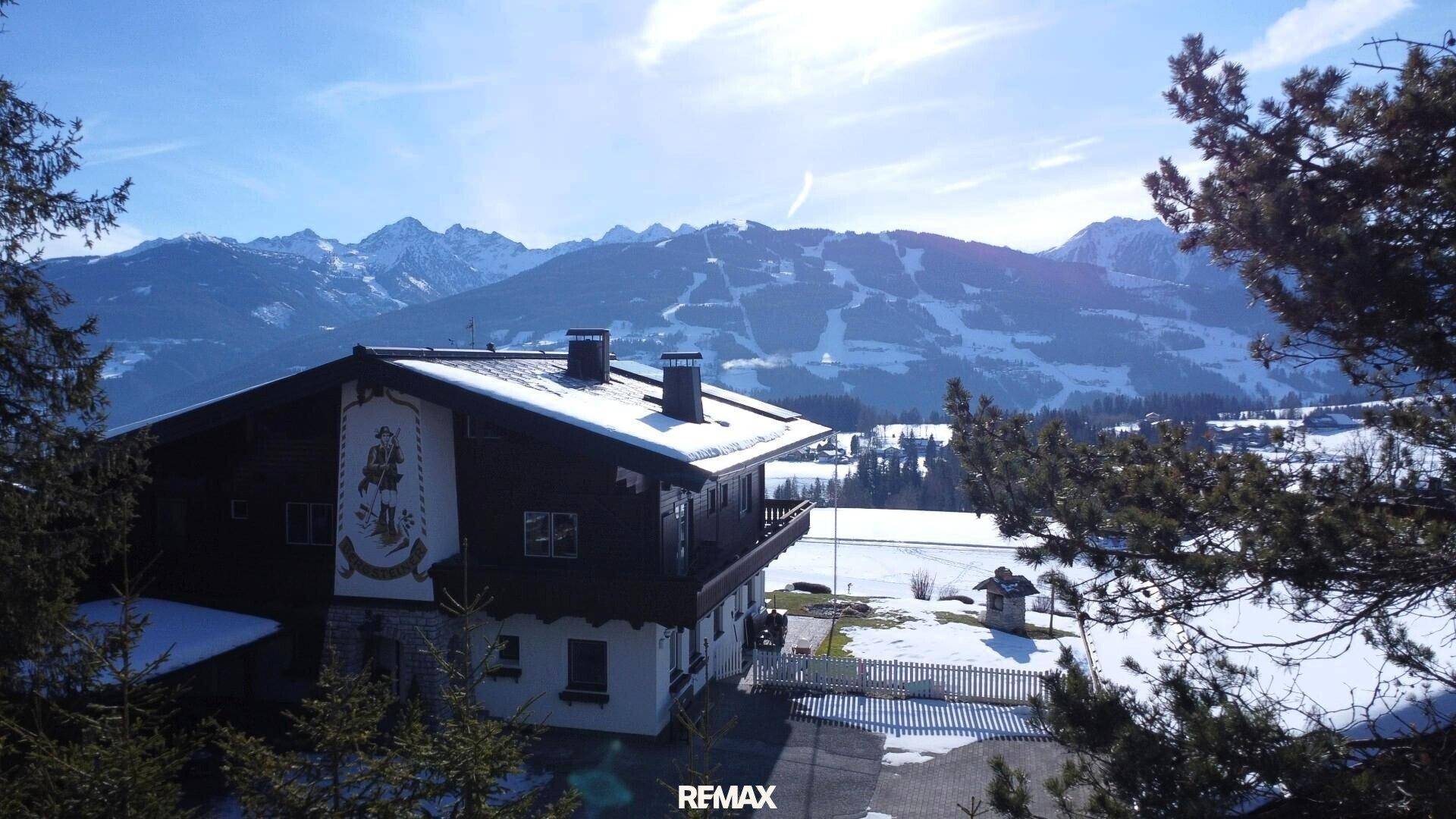 Traditionelles Chalet-Gebäude mit schneebedecktem Dach und beeindruckendem Bergpanorama im Hintergrund.