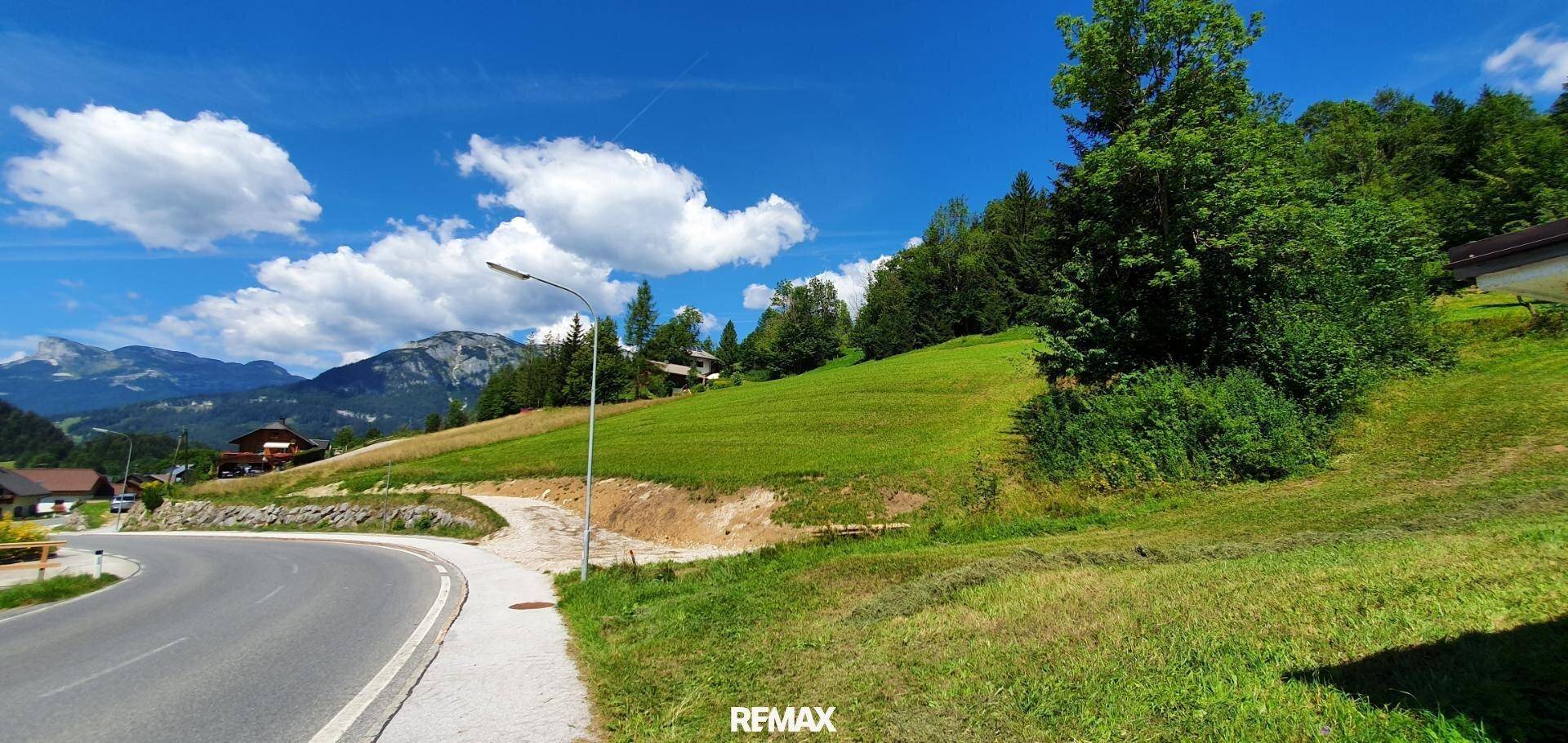 Landstraße mit Gehweg und Laterne, umgeben von grünen Hügeln und fernen Bergen.