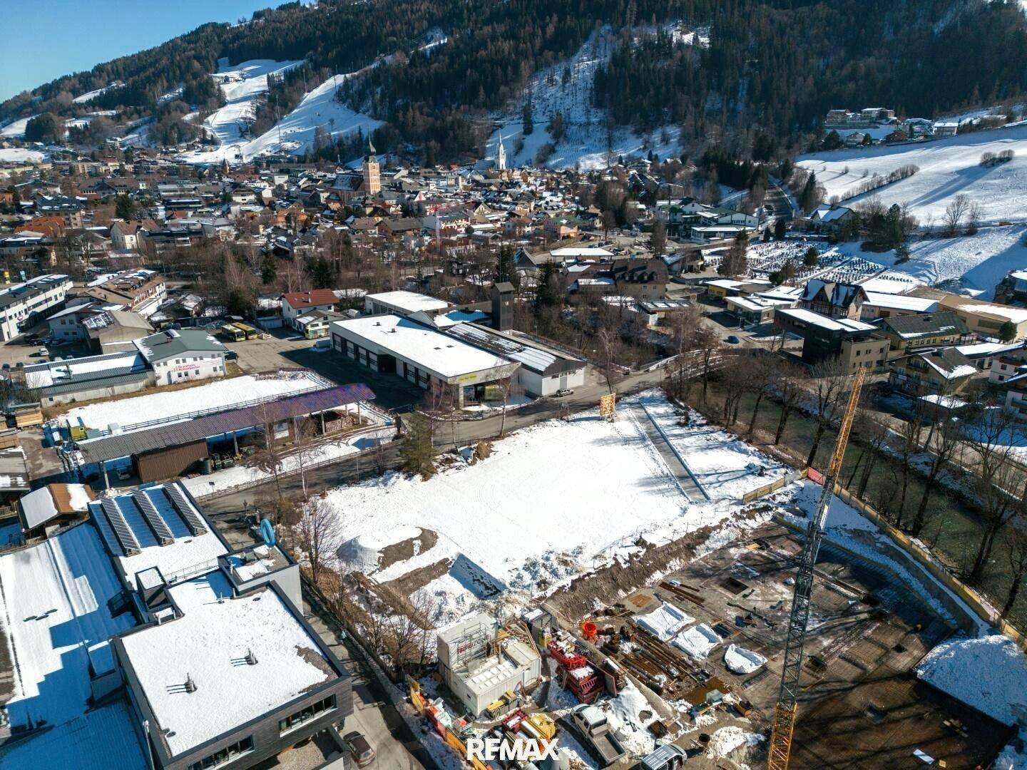 Winterliche Luftaufnahme einer Baustelle und der umliegenden Stadt vor schneebedeckten Bergen.