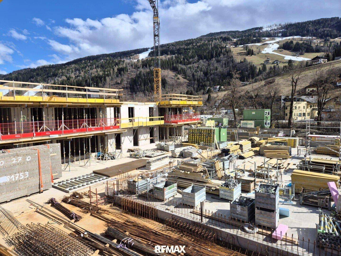 Baustelle mit im Bau befindlichen Gebäuden und Baumaterialien vor verschneiter Bergkulisse.