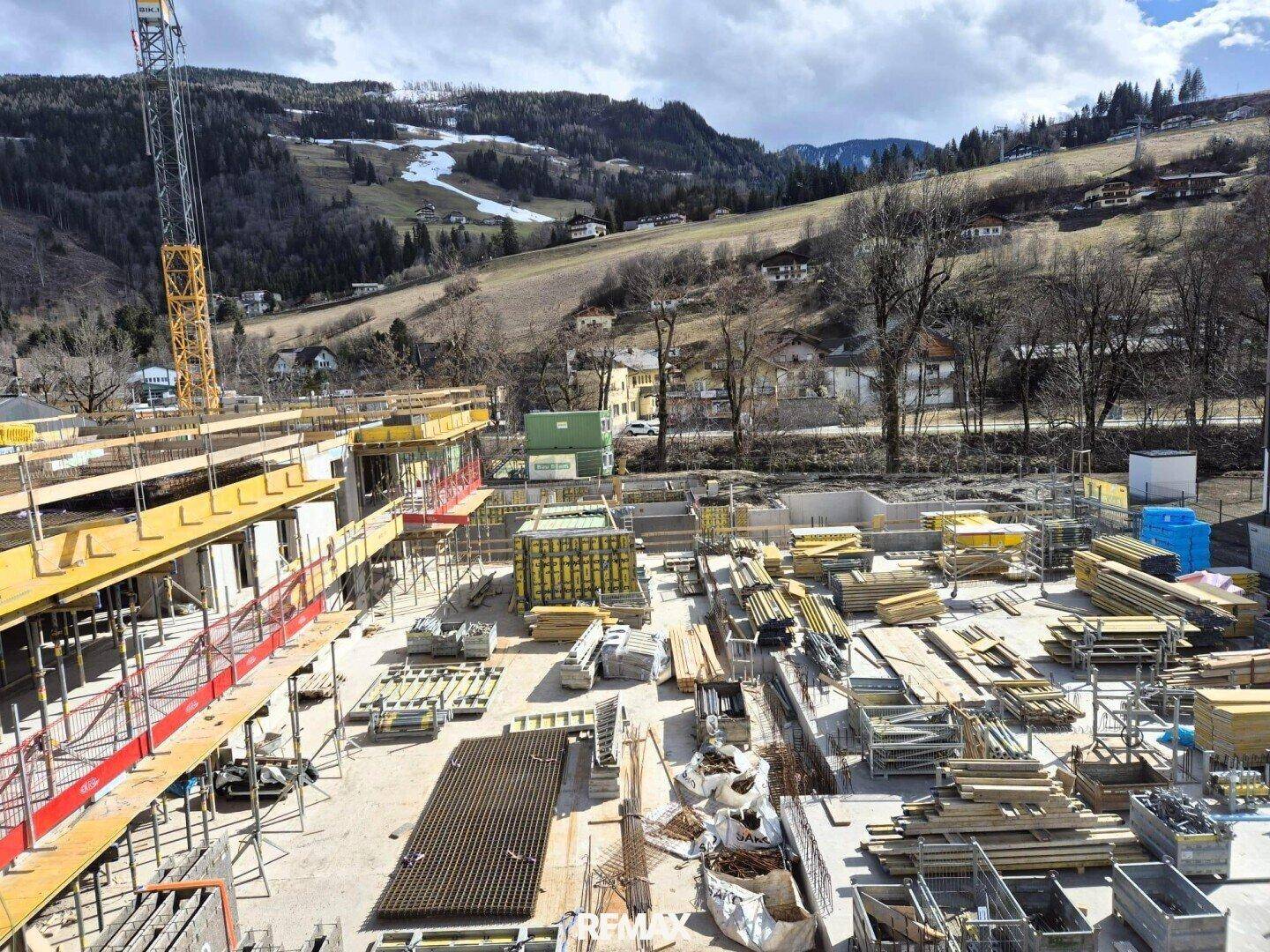 Baustelle mit Rohbauten und gelagerten Materialien, umgeben von winterlicher Berglandschaft.