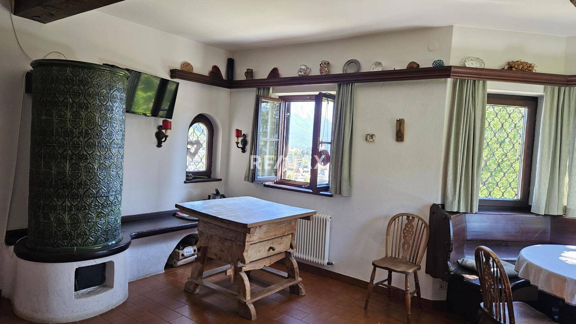 Traditioneller Essbereich mit Kachelofen, Holztisch und Fenstern mit Blick ins Grüne.