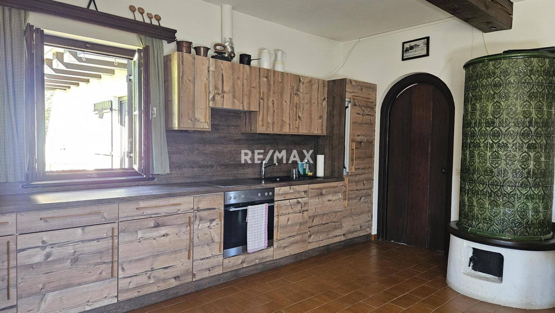 Helle Küche mit Holzfronten, Kachelofen und Fenster, das viel Tageslicht hereinlässt.