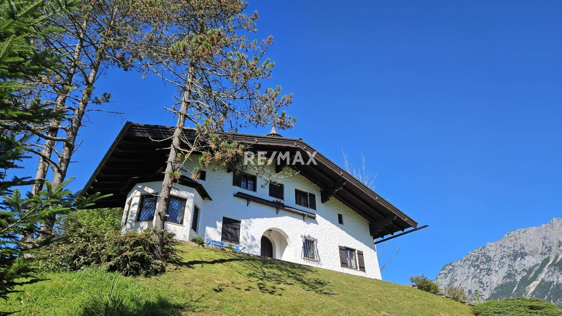 Traditionelles Haus auf einem Hügel mit Bäumen und Bergen im Hintergrund unter blauem Himmel.