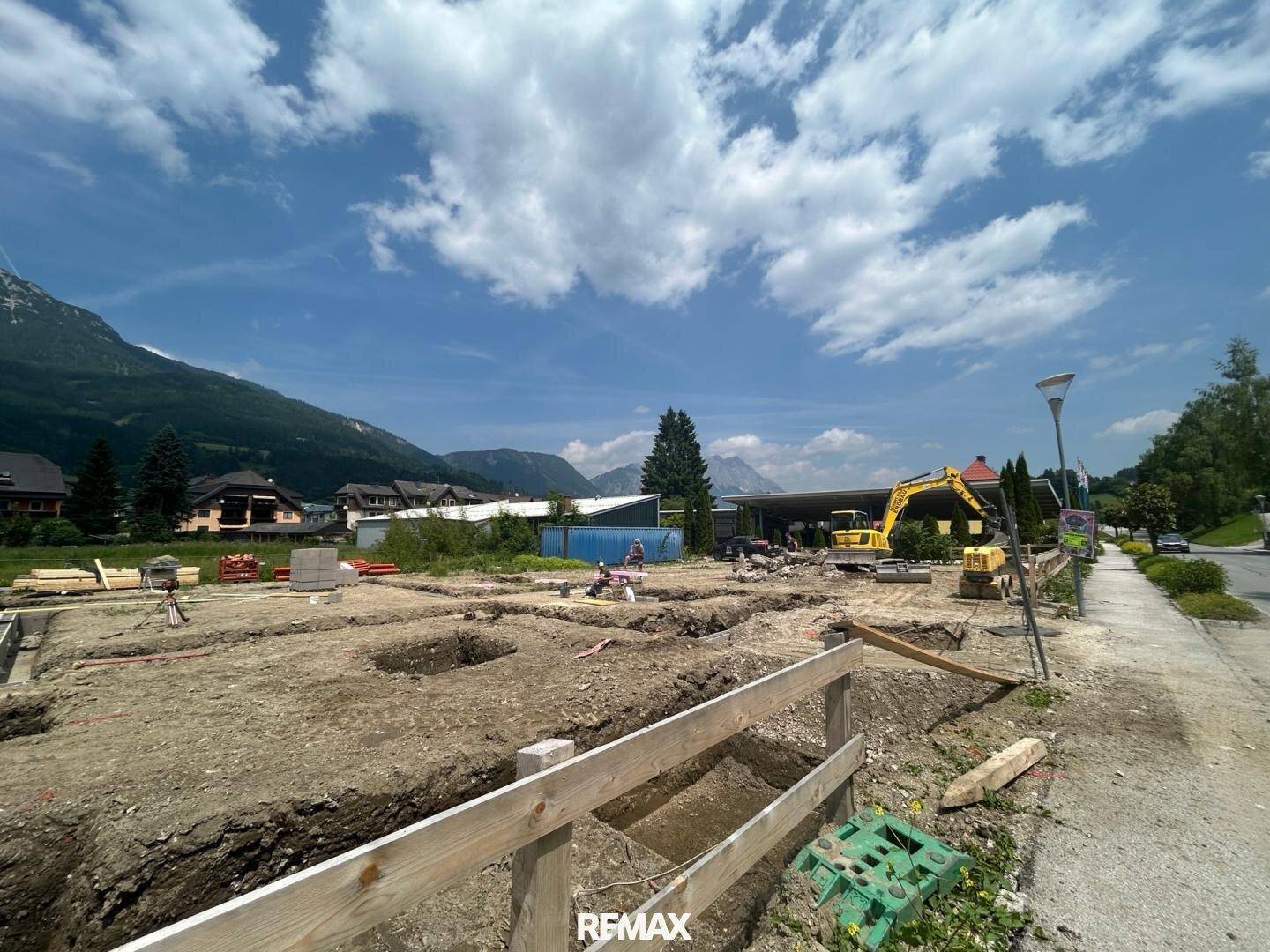 Baustelle mit ausgehobenen Fundamenten und Baugeräten vor bergiger Landschaft.
