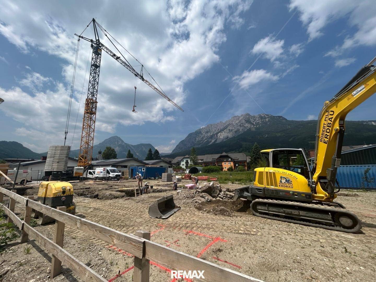 Baustelle mit Bagger und Kran vor einer beeindruckenden Bergkulisse unter blauem Himmel.