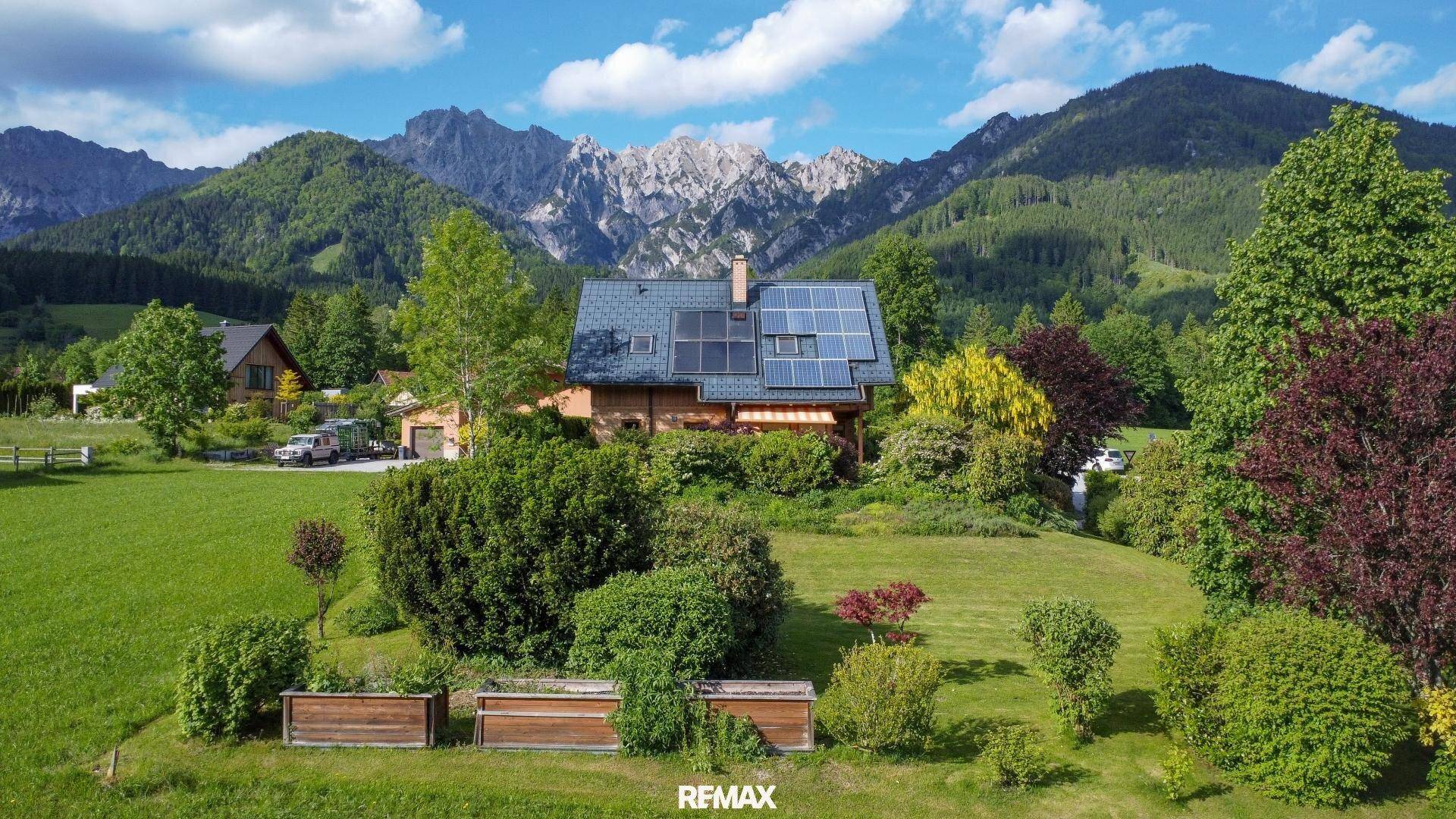 Luftaufnahme des Holzhauses mit Solaranlage, umgeben von grünen Wiesen und Bergen.