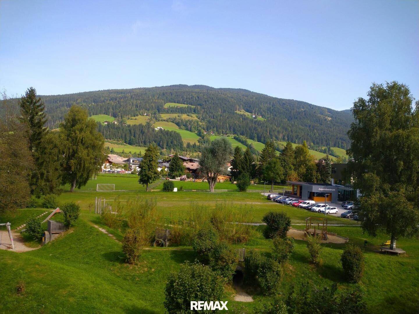 Panoramablick auf eine weite grüne Berglandschaft mit einem Tal und einigen Gebäuden in der Ferne.