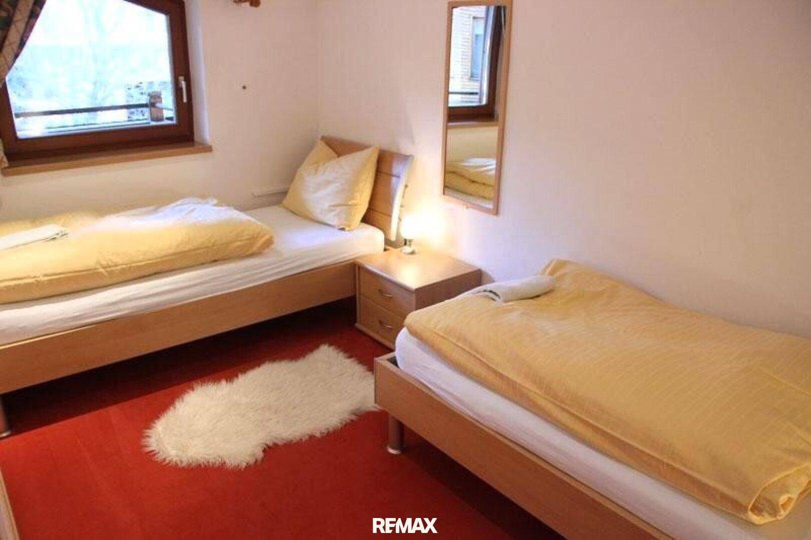 Helles Schlafzimmer mit zwei Einzelbetten und rotem Bodenbelag, ausgestattet mit Holzmöbeln.