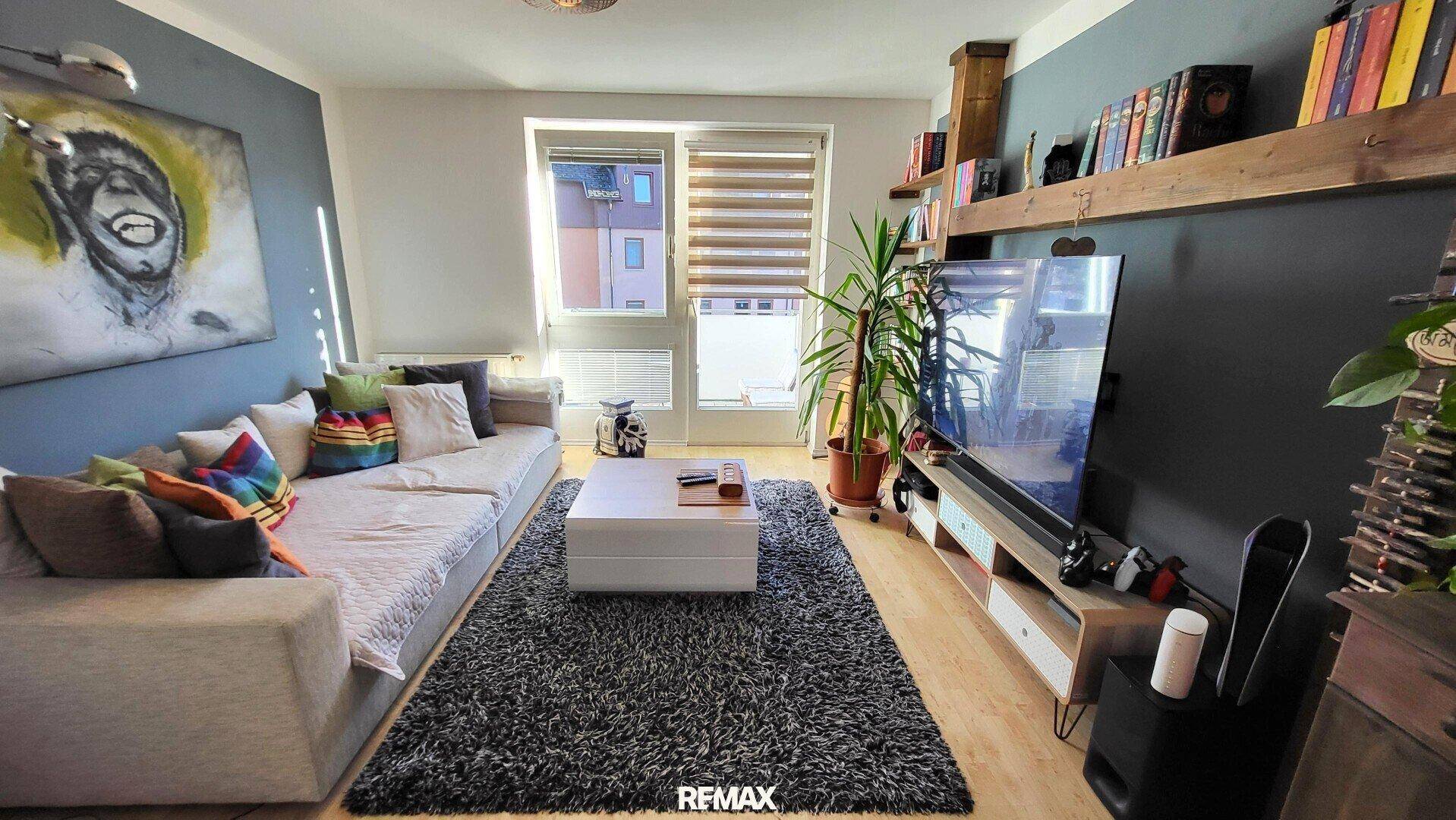 Helles Wohnzimmer mit Sofa, Fernseher, Bücherregal und großen Fenstern.