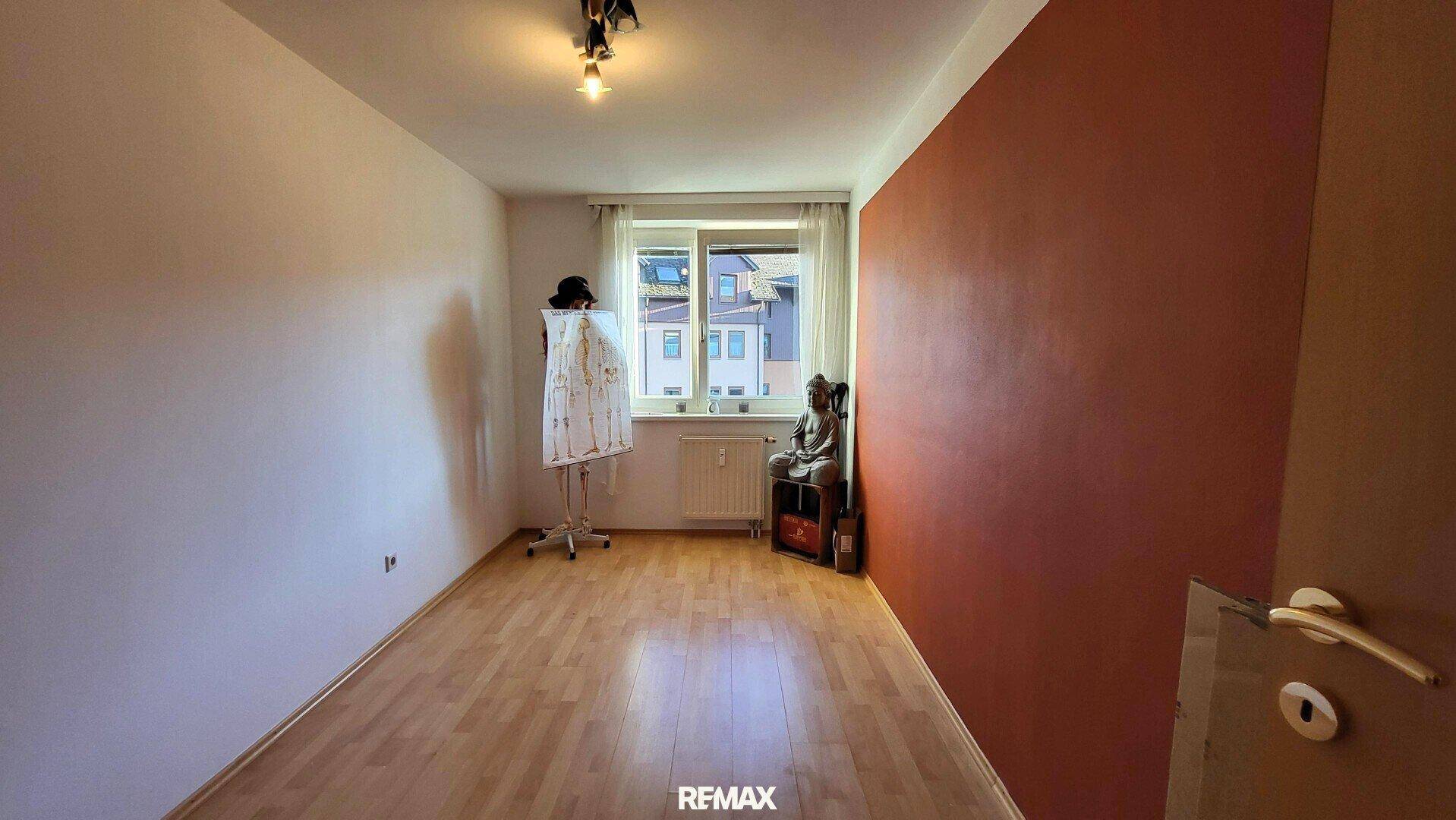 Leeres Schlafzimmer mit roter Akzentwand, Parkettboden und Fenster.