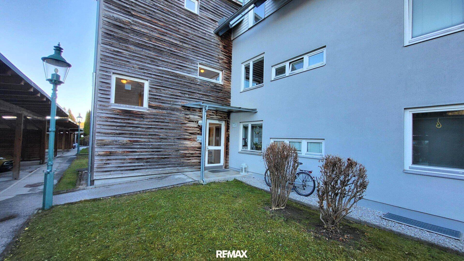 Eingangsbereich des Gebäudes mit Holzfassade, Grünfläche und Carport.