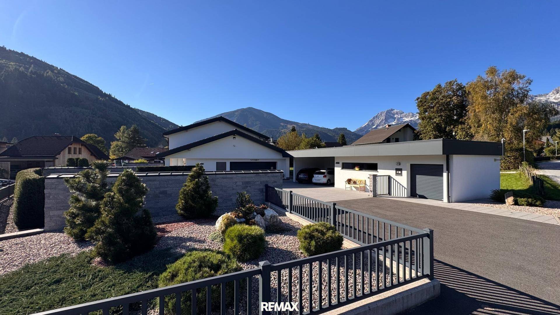 Ansicht der modernen Villa mit Zufahrt, Carport und gepflegtem Vorgarten vor alpiner Kulisse.