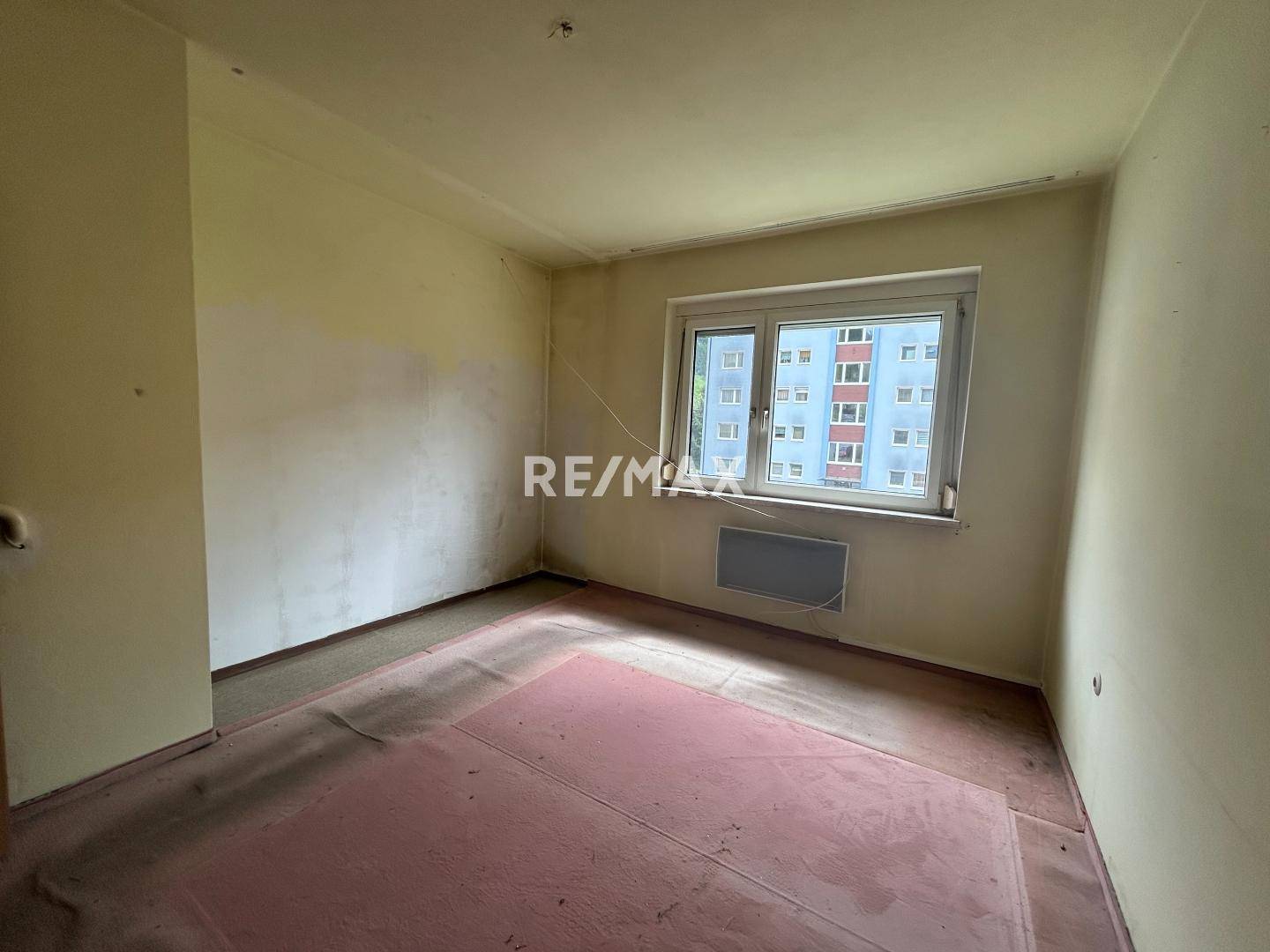 Leeres Zimmer mit Teppichboden und einem Fenster, das auf die gegenüberliegenden Gebäude blickt.