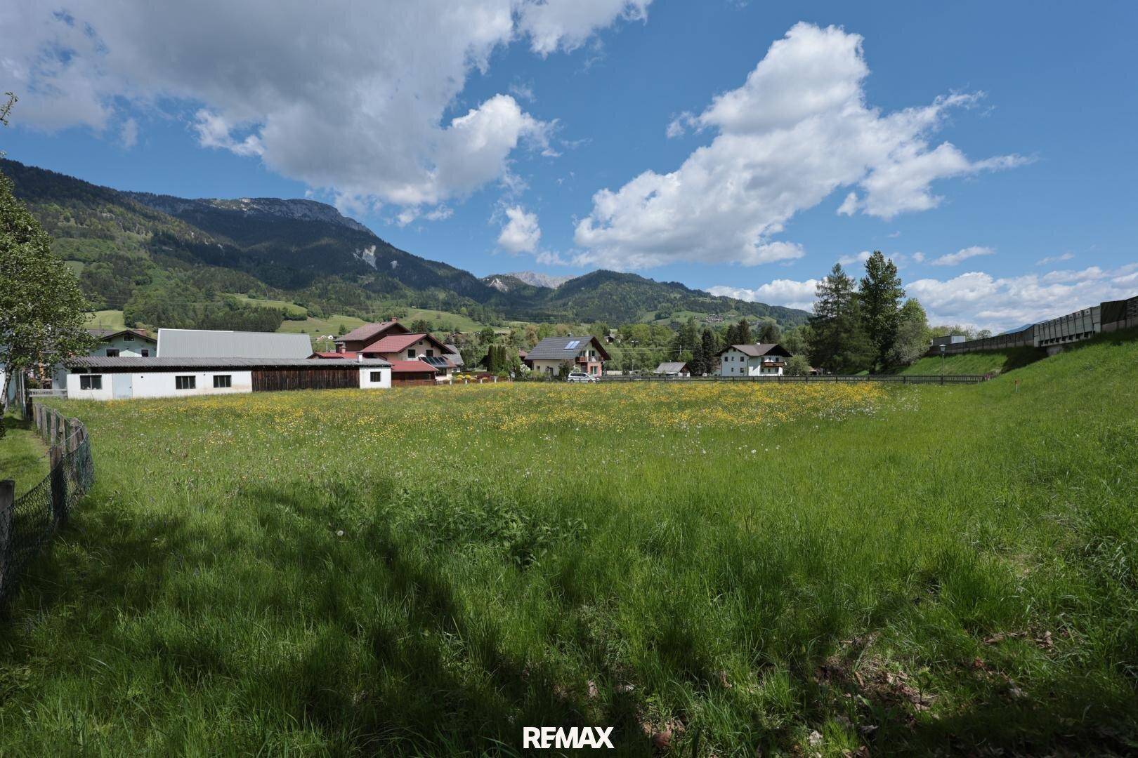 Weitläufige grüne Wiese mit Blick auf die umliegende Berglandschaft und vereinzelte Häuser.