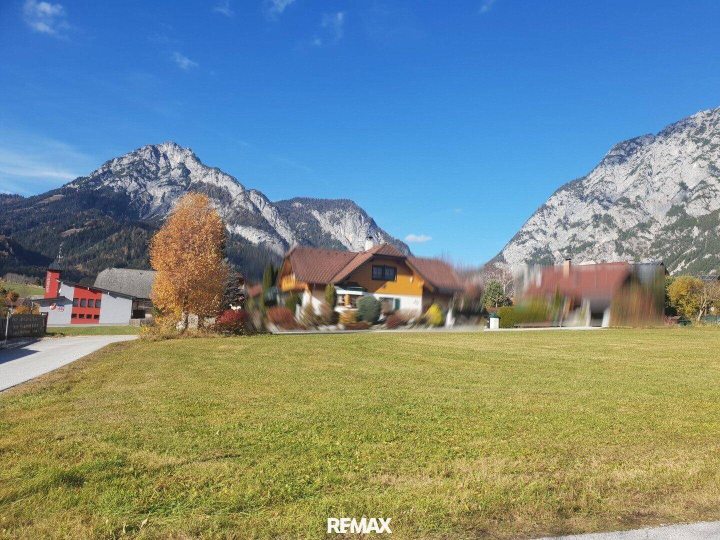 Sonnige Wiesenlandschaft mit Häusern, zwei Berggipfeln und einer Straße im Vordergrund.