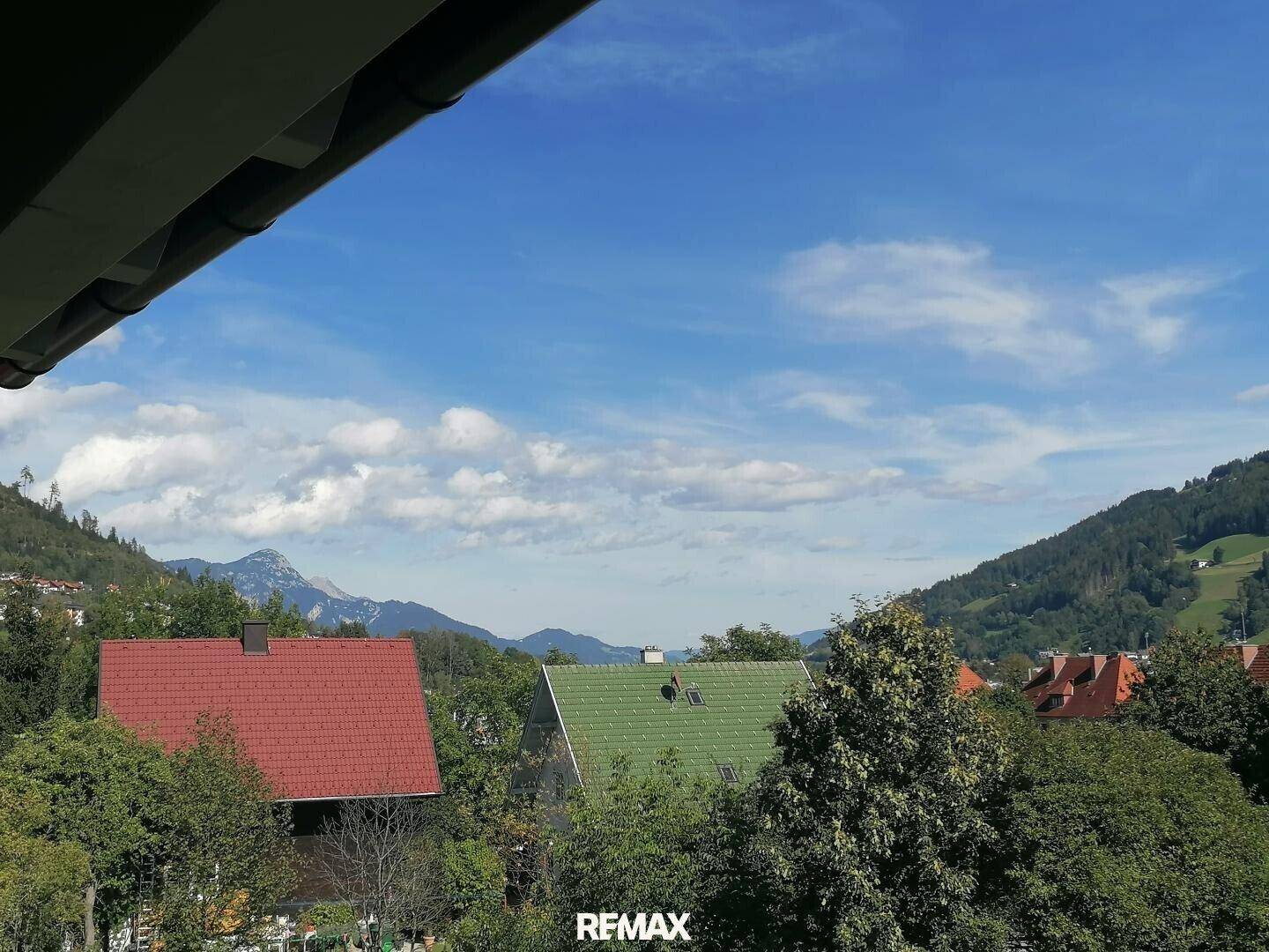 Panoramablick über Dächer und grüne Hügel mit Bergen im Hintergrund unter blauem Himmel.