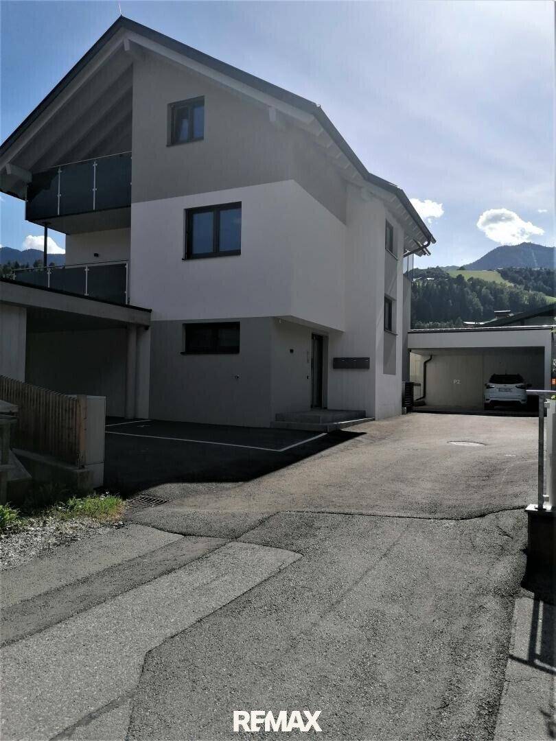 Gesamtansicht des modernen Gebäudes mit Eingangsbereich, Carport und Balkonen vor Bergkulisse.