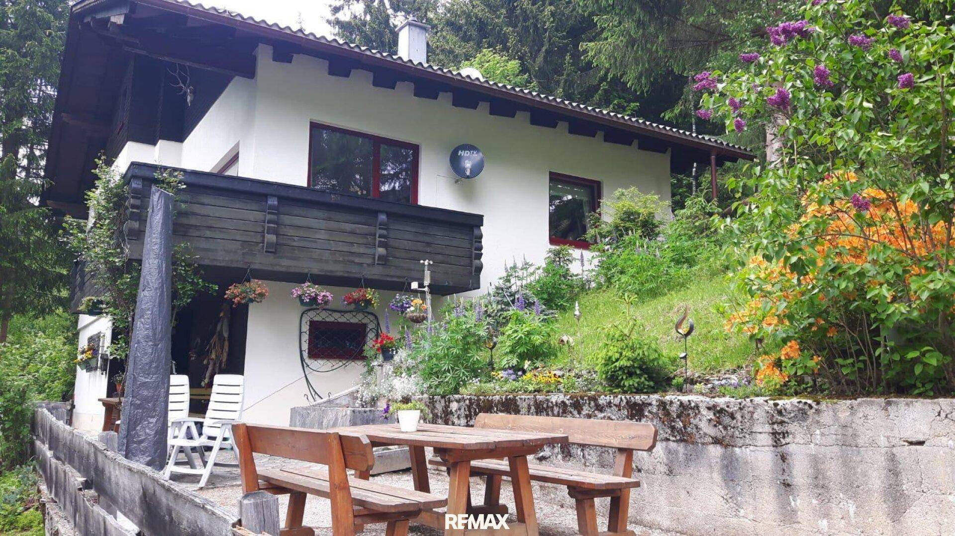 Traditionelles Haus mit Balkon und blühenden Blumen, umgeben von üppiger Vegetation und einem sonnigen Garten.