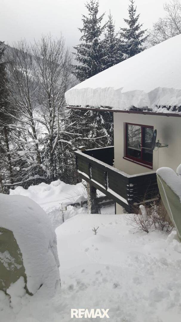 Hausfassade im Winter mit schneebedecktem Dach und Balkon, umgeben von verschneiten Bäumen.