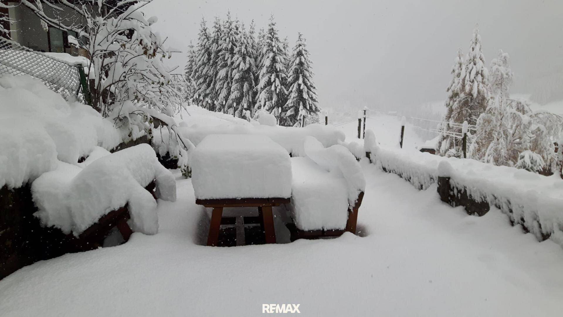 Verschneiter Außenbereich mit Holztisch und Bänken, umgeben von einer winterlichen Landschaft.