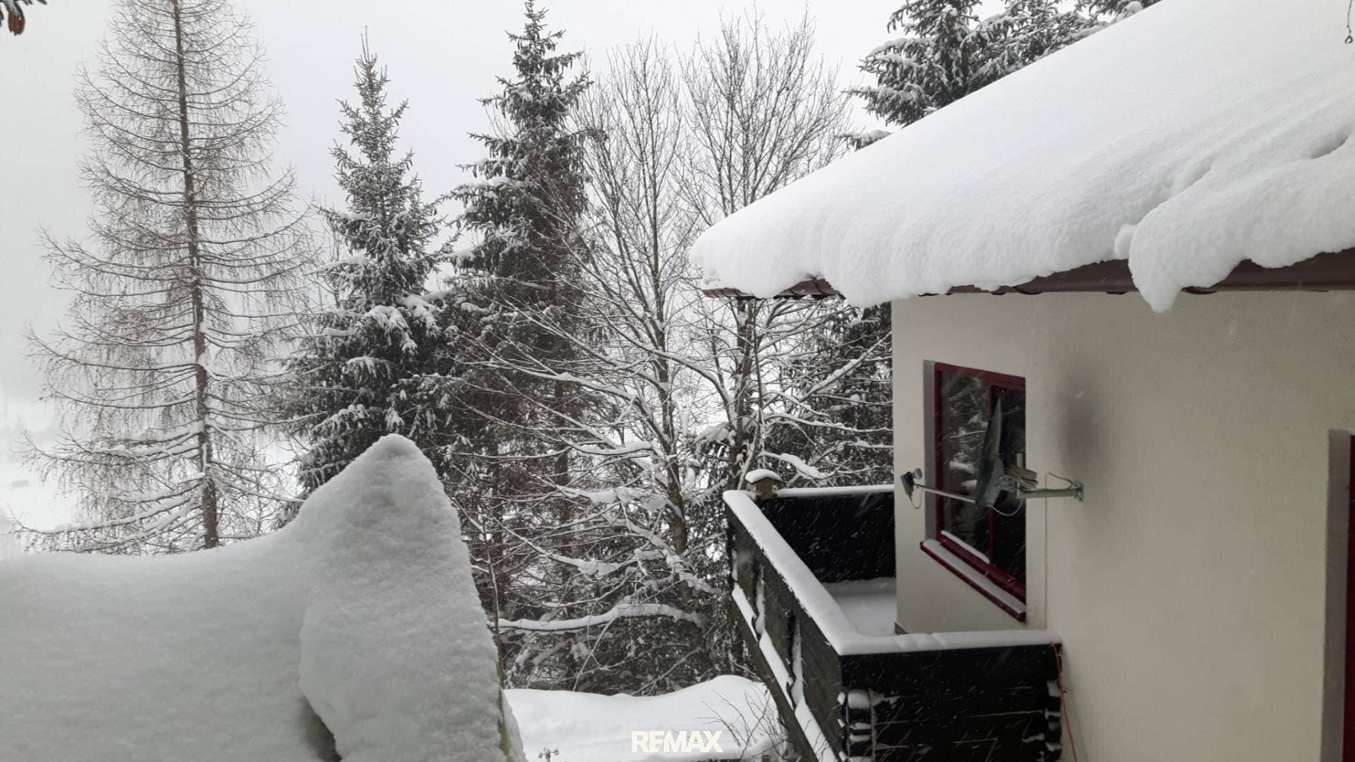 Hausfassade im Winter mit schneebedecktem Dach und Balkon, umgeben von verschneiten Bäumen.