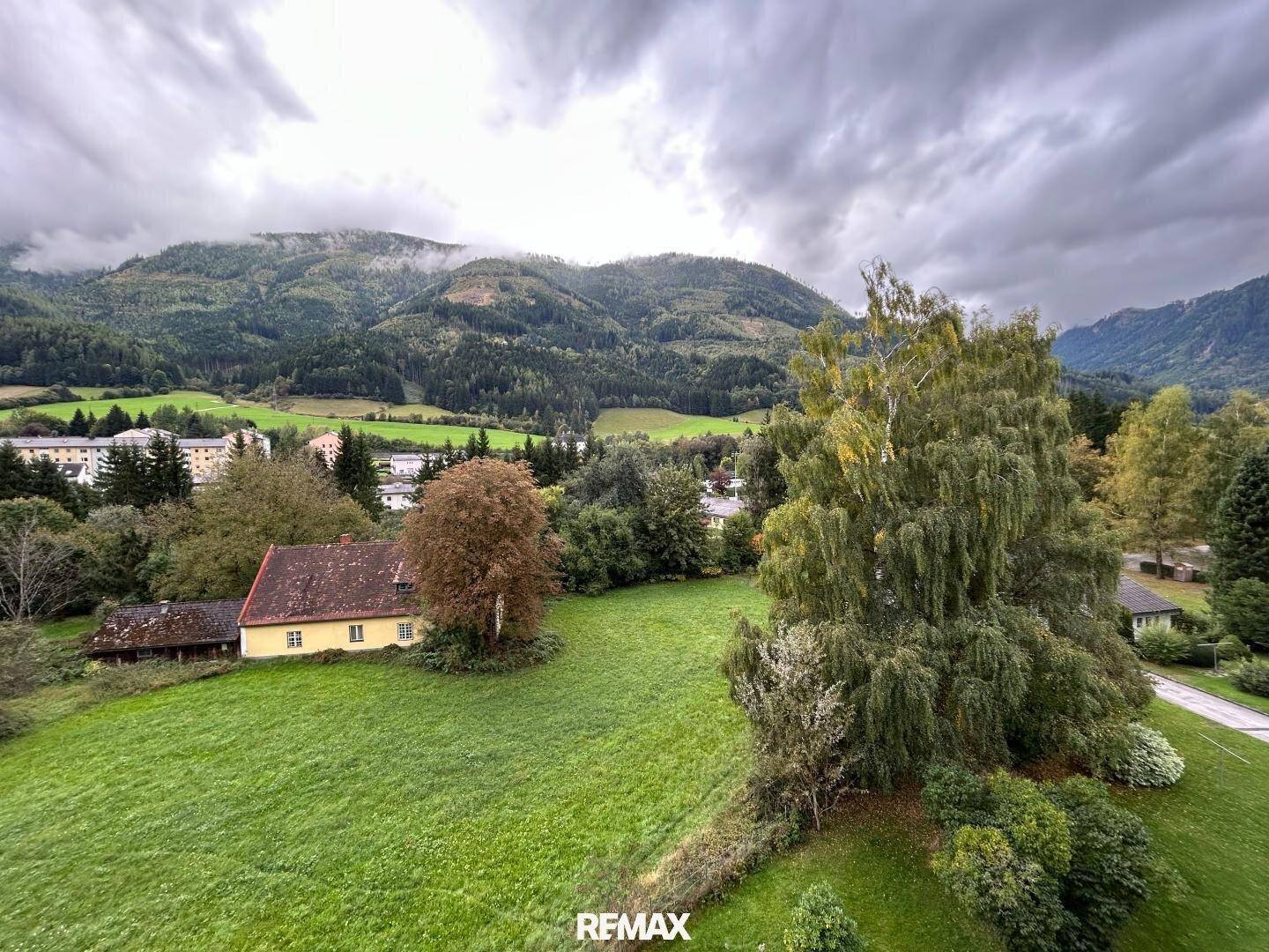 Weitläufiger Blick auf grüne Wiesen und bewaldete Berge unter einem bewölkten Himmel.