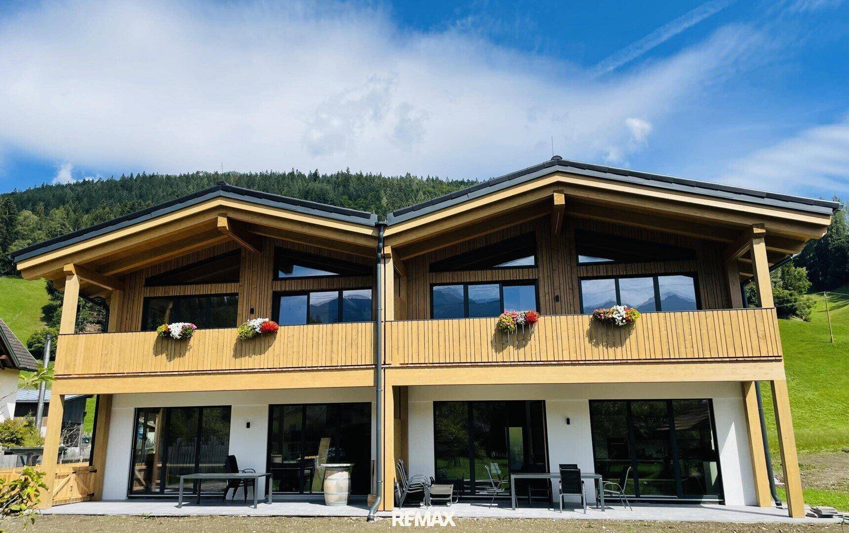 Modernes Chalet mit Holzfassade, großen Fenstern und Terrasse vor grüner Bergkulisse.
