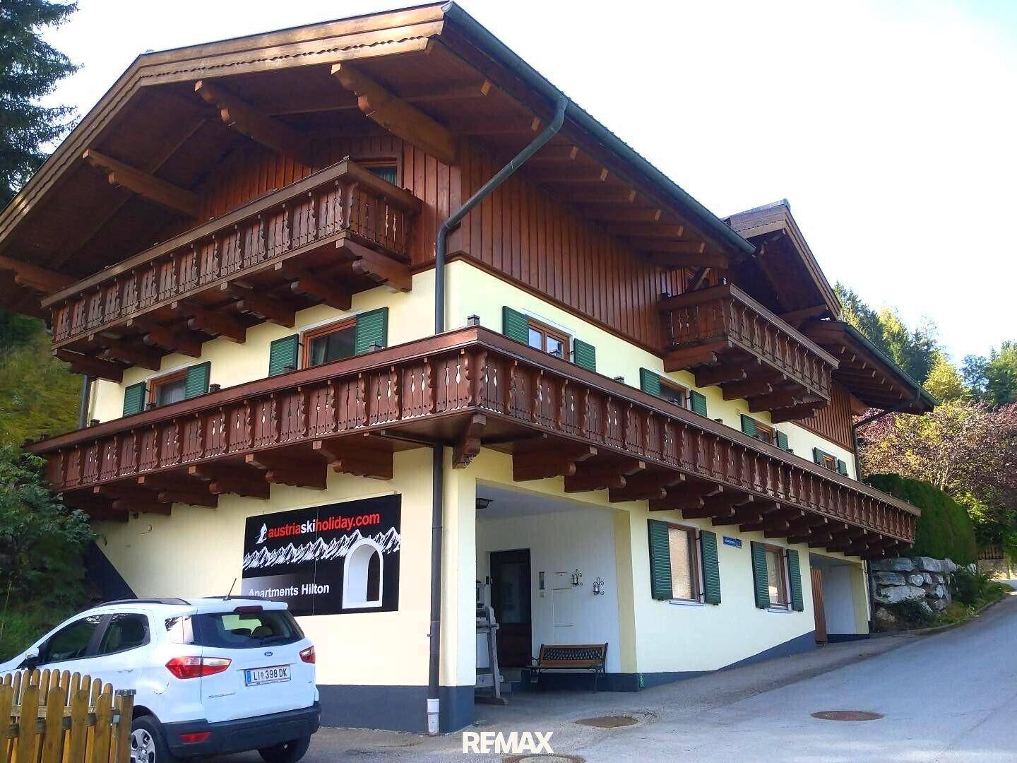 Traditionelles Gebäude im alpinen Stil mit mehreren Holzbalkonen und einer hellgelben Fassade an einem Hang.