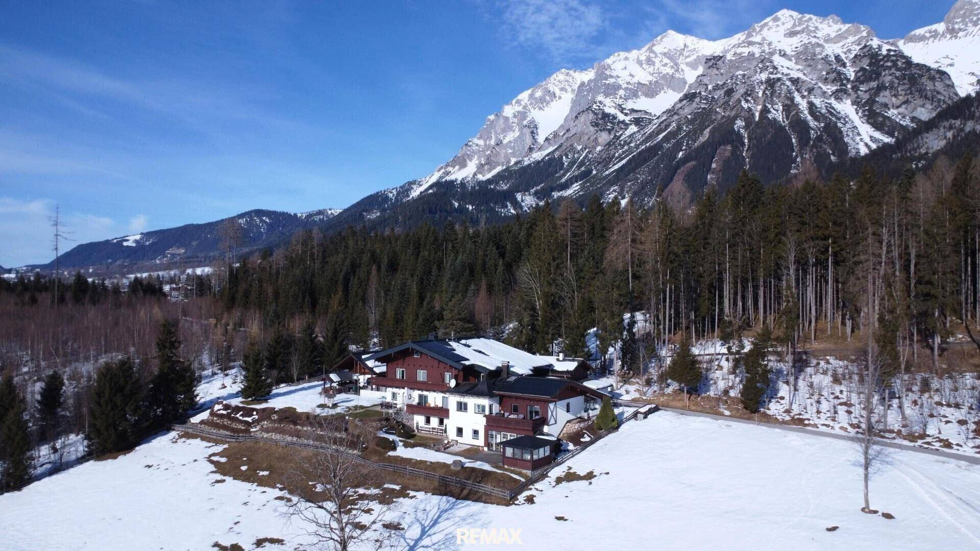 Weitläufiges Anwesen in einer verschneiten Waldlichtung mit majestätischen Bergen unter blauem Himmel.