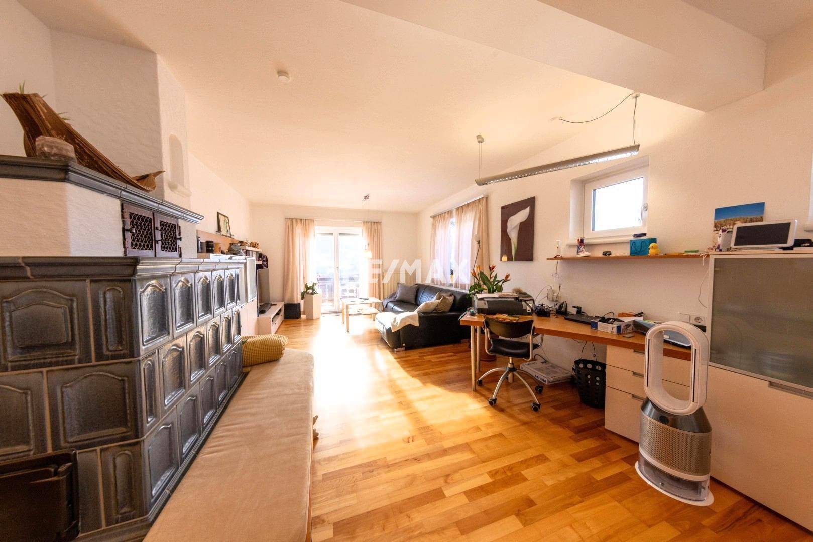 Helles Wohnzimmer mit Holzboden, Kachelofen und Zugang zu einem Balkon, gemütlich eingerichtet.