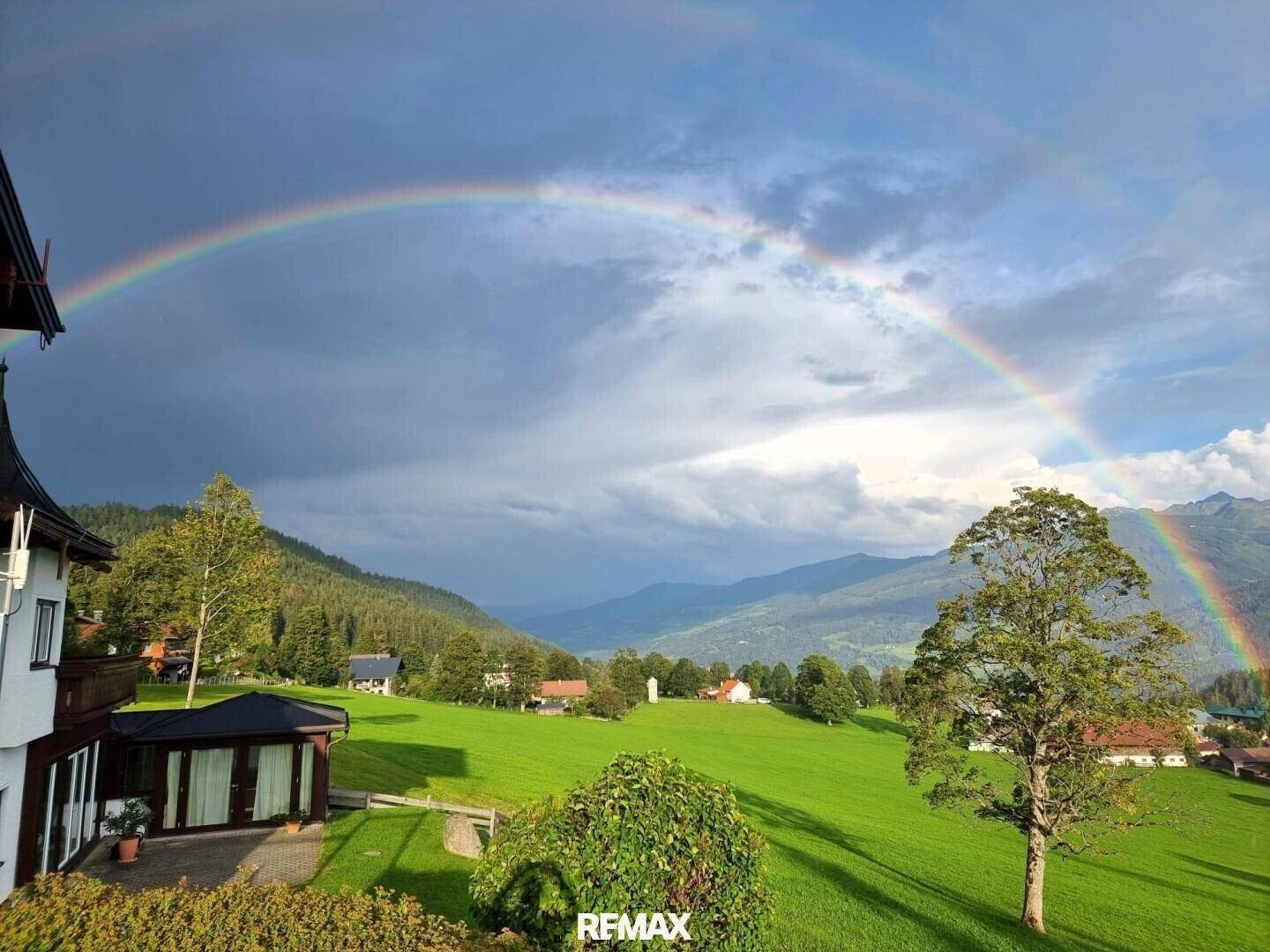 Haus in idyllischer Landschaft mit weiten grünen Wiesen und einem beeindruckenden Regenbogen über den Bergen.