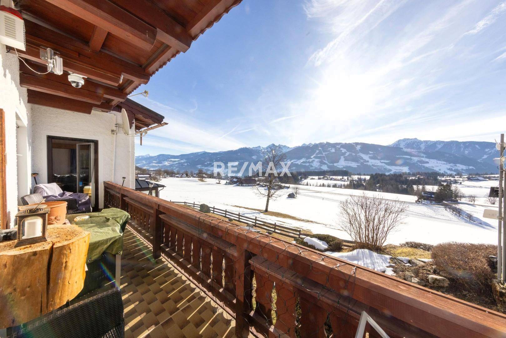 Sonniger Holzbalkon mit traditionellem Geländer und weitem Panoramablick auf die verschneite Berglandschaft.