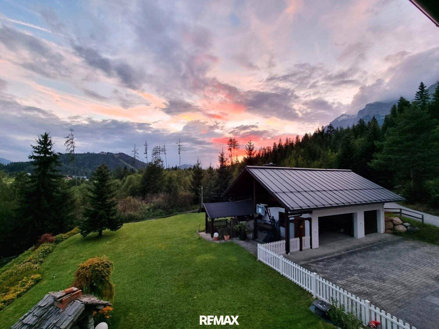 Malerisches Haus mit Garage und grüner Wiese vor einer Bergkulisse bei Sonnenuntergang.