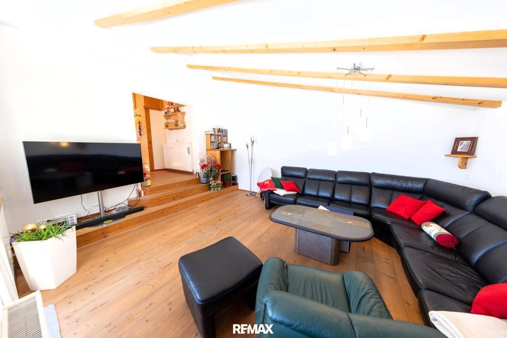 Geräumiges Wohnzimmer mit schwarzem Ecksofa, Holzboden und dekorativen Holzbalken an der Decke.