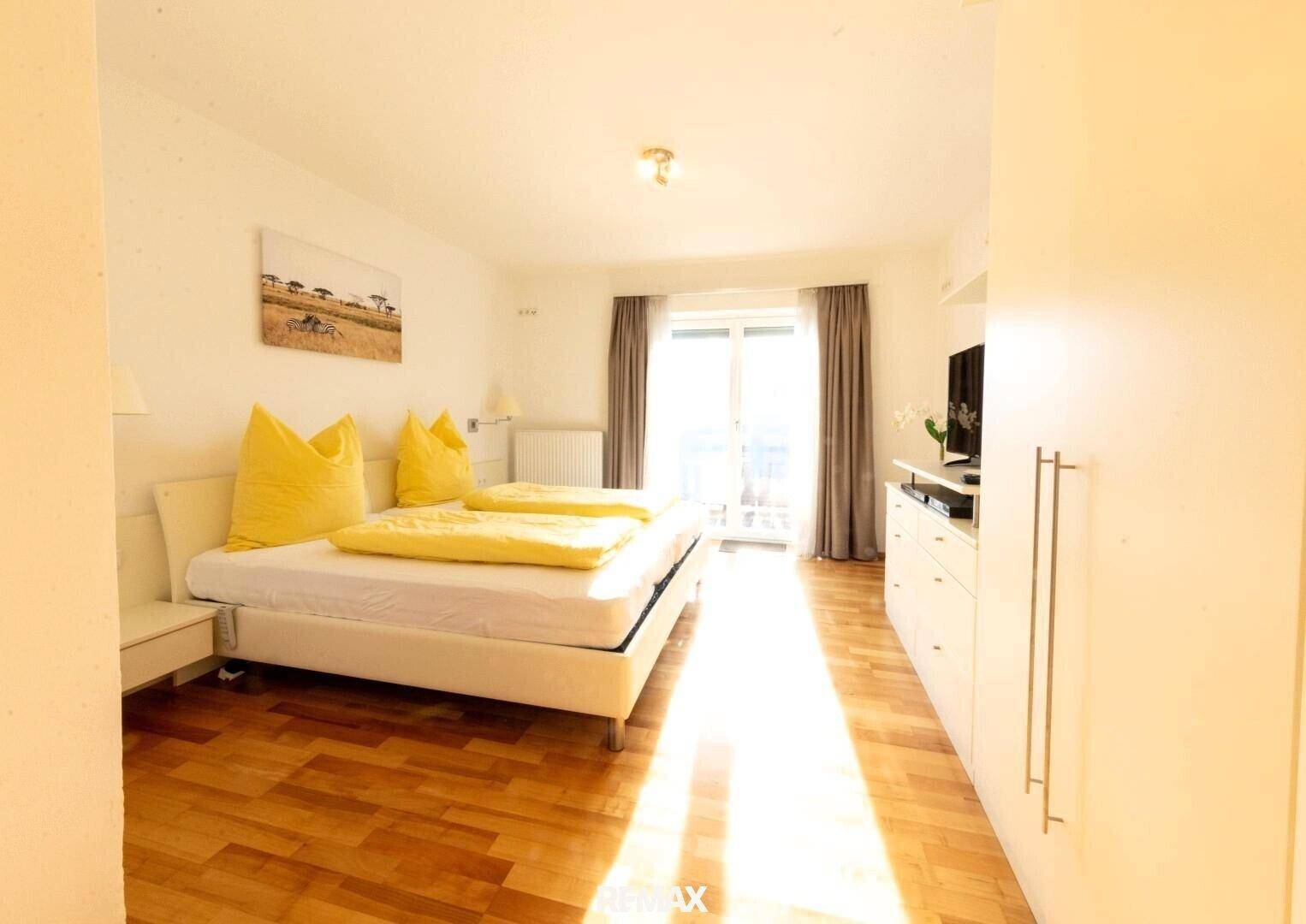 Helles Schlafzimmer mit Holzboden, weißem Bett und Zugang zu einem Balkon, durchflutet von Sonnenlicht.