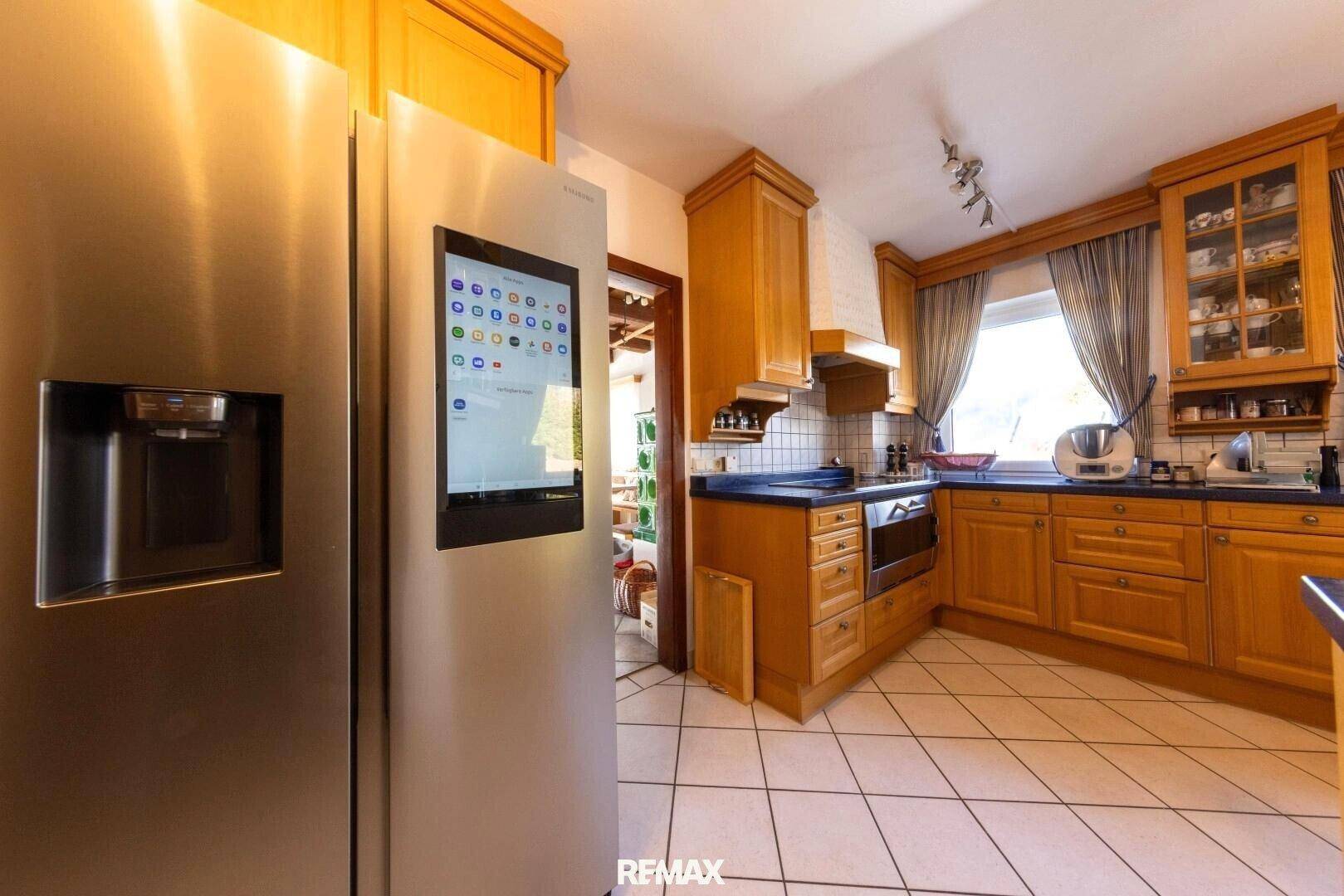 Funktionale Küche mit Holzschränken, Fliesenboden und einem modernen Kühlschrank mit integriertem Display.