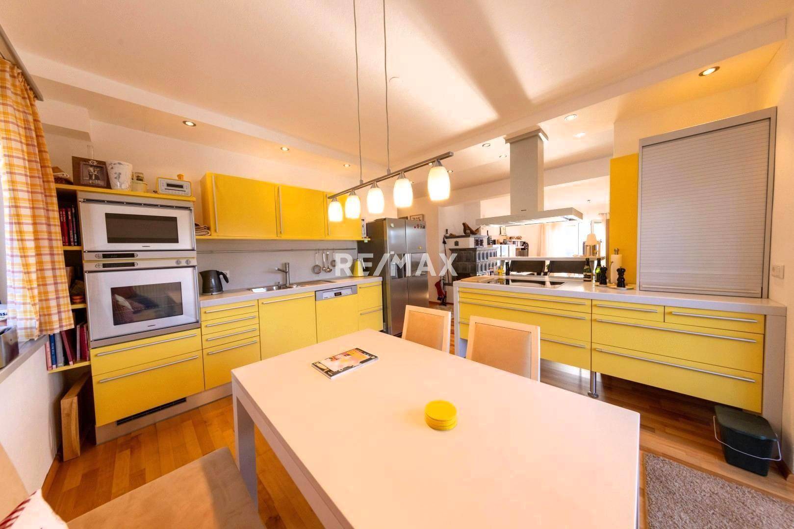 Moderne gelbe Küche mit Essbereich, Kochinsel und hochwertigen Einbaugeräten, sehr hell.