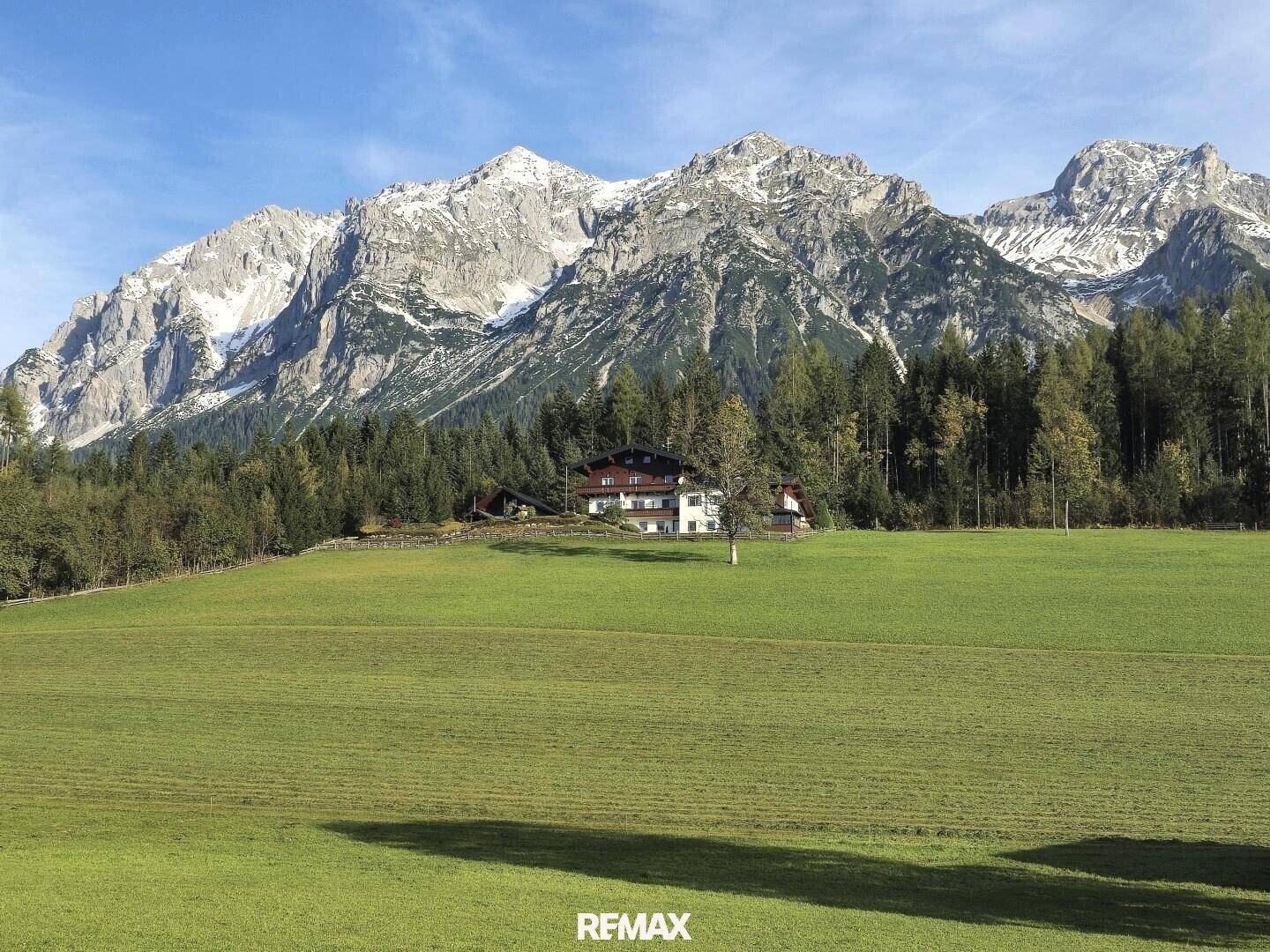 Traditionelles Haus inmitten einer weiten grünen Wiese mit majestätischen schneebedeckten Bergen im Hintergrund.