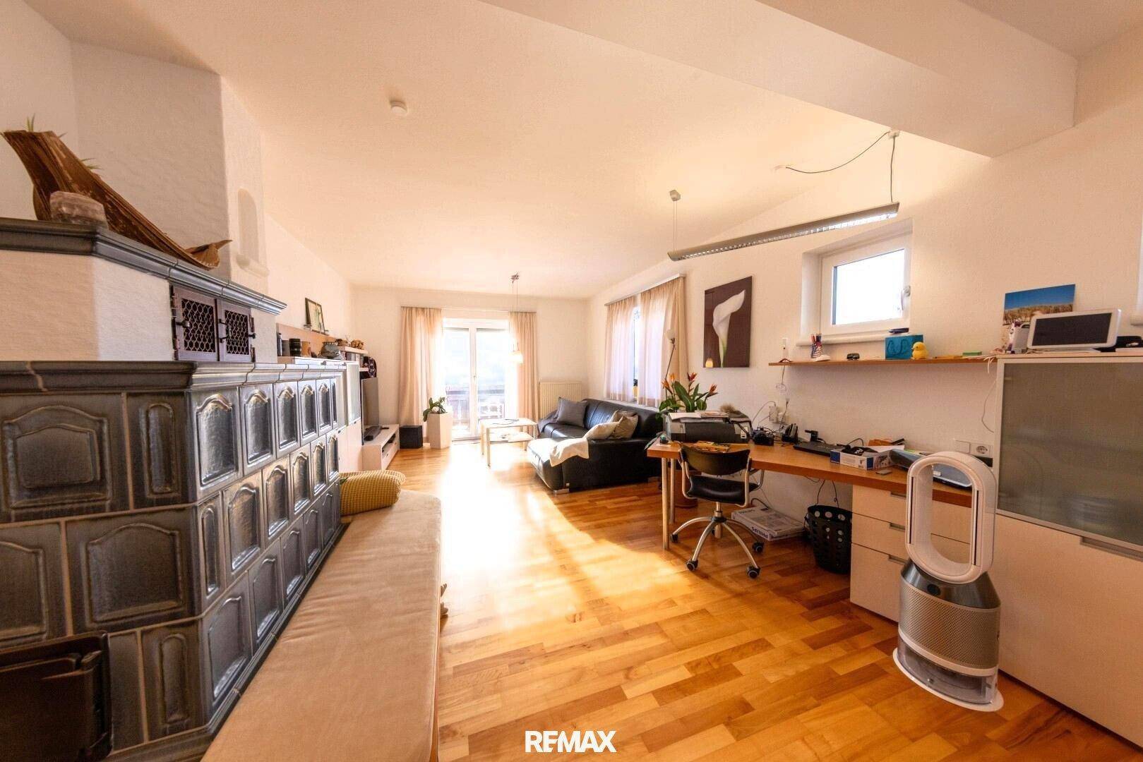 Gemütliches Wohnzimmer mit traditionellem Kachelofen, Sofa, Schreibtisch und sonnigem Balkonzugang.