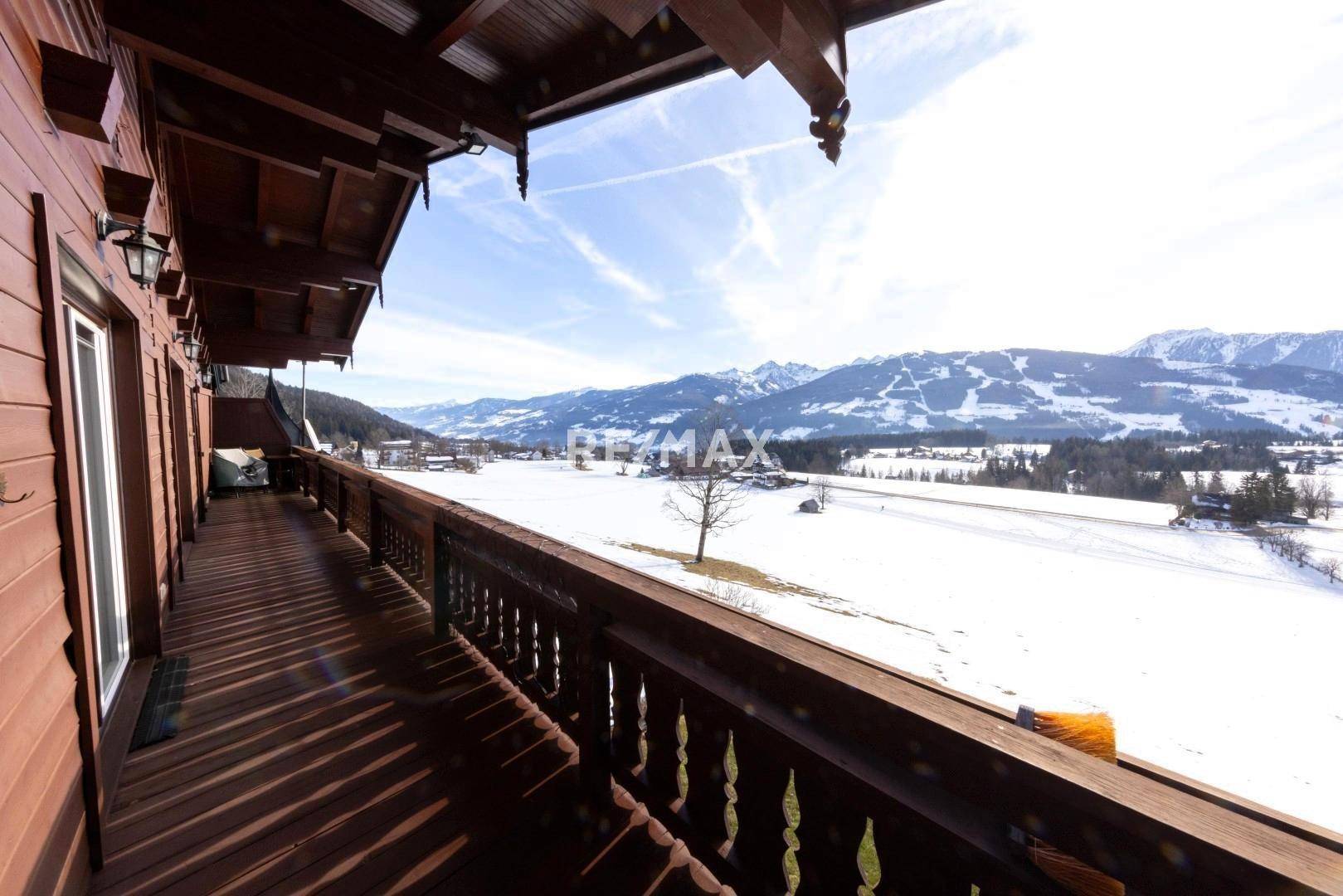 Sonniger Holzbalkon mit traditionellem Geländer und weitem Panoramablick auf die verschneite Bergwelt.