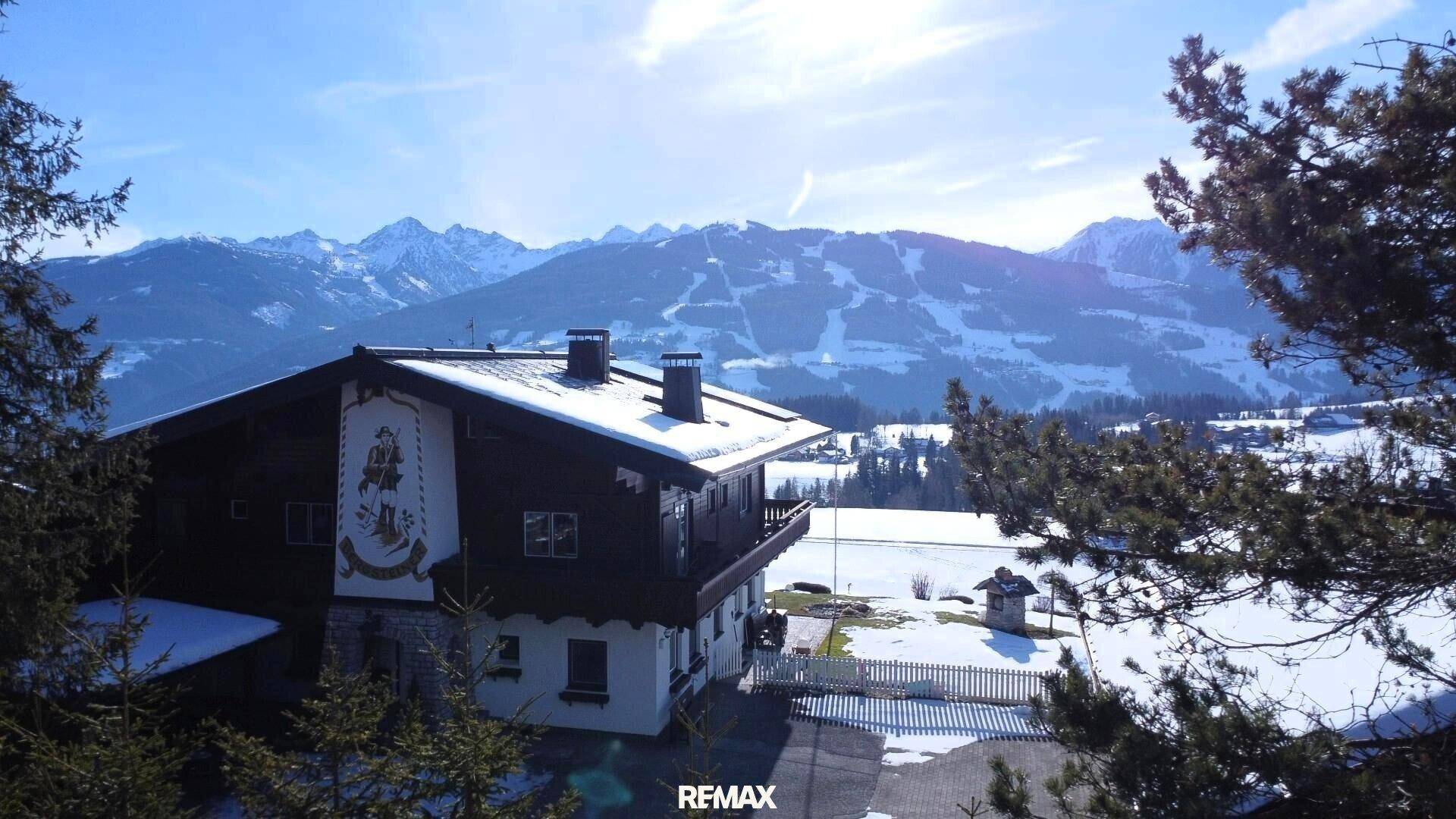 Traditionelles Haus mit alpiner Fassadenmalerei in verschneiter Umgebung und Blick auf Skipisten.