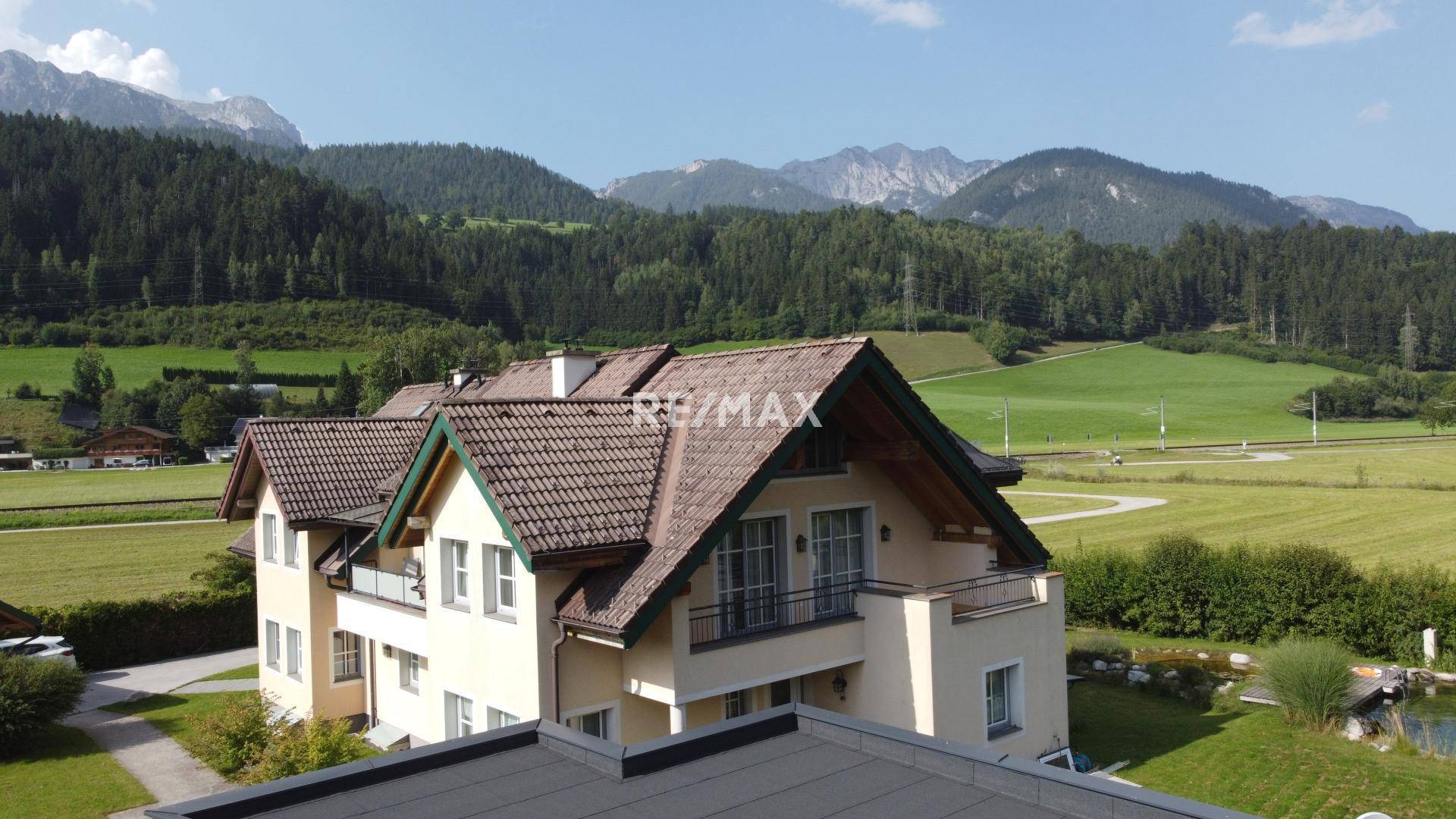 Panoramablick auf das Haus mit Balkonen, umgeben von weiten Feldern und majestätischen Bergen.