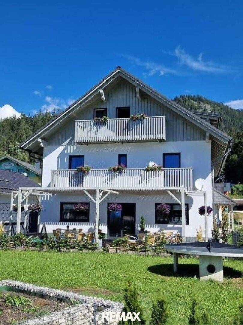 Ansicht des weißen Hauses mit Balkonen und einer Terrasse im Gartenbereich, umgeben von Natur.